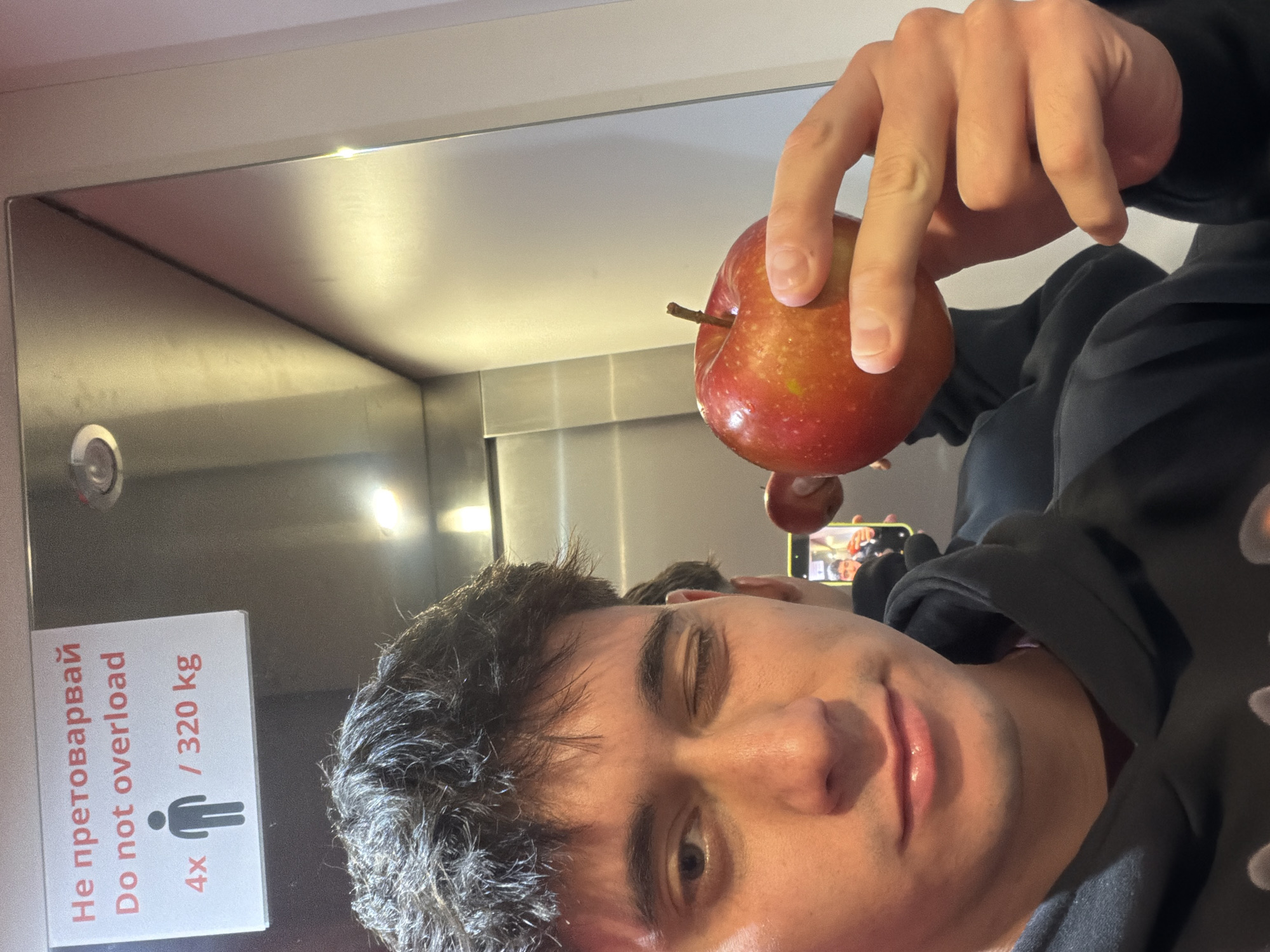 A selfie taken in an elevator. A fair-skinned person with dark hair is holding a red apple in their right hand. The elevator walls are stainless steel. A sign indicating the elevator capacity is visible, stating "Do not overload" and "4x / 320 kg" in English and Bulgarian. The photograph was likely taken in Sofia, Bulgaria, as the Bulgarian language is present on the elevator sign.