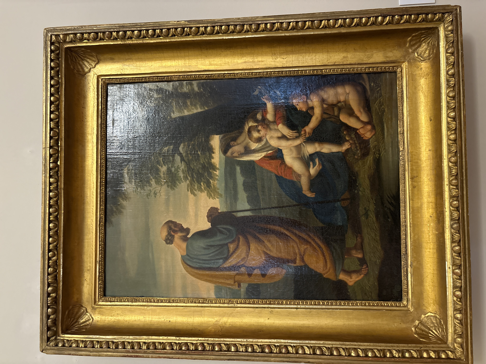 The image shows a framed, rectangular painting. The painting depicts a scene with an older, bearded man in a brown robe and blue shirt, carrying a walking stick, walking towards the left side of the frame. A woman wearing a blue robe and red garment is holding a baby, being reached for by two nude children. The background is a landscape with trees and sky. The painting is framed in a heavily ornate, gold-colored frame. The image was taken in Saint Petersburg, Russia, although the location in that city is unknown.