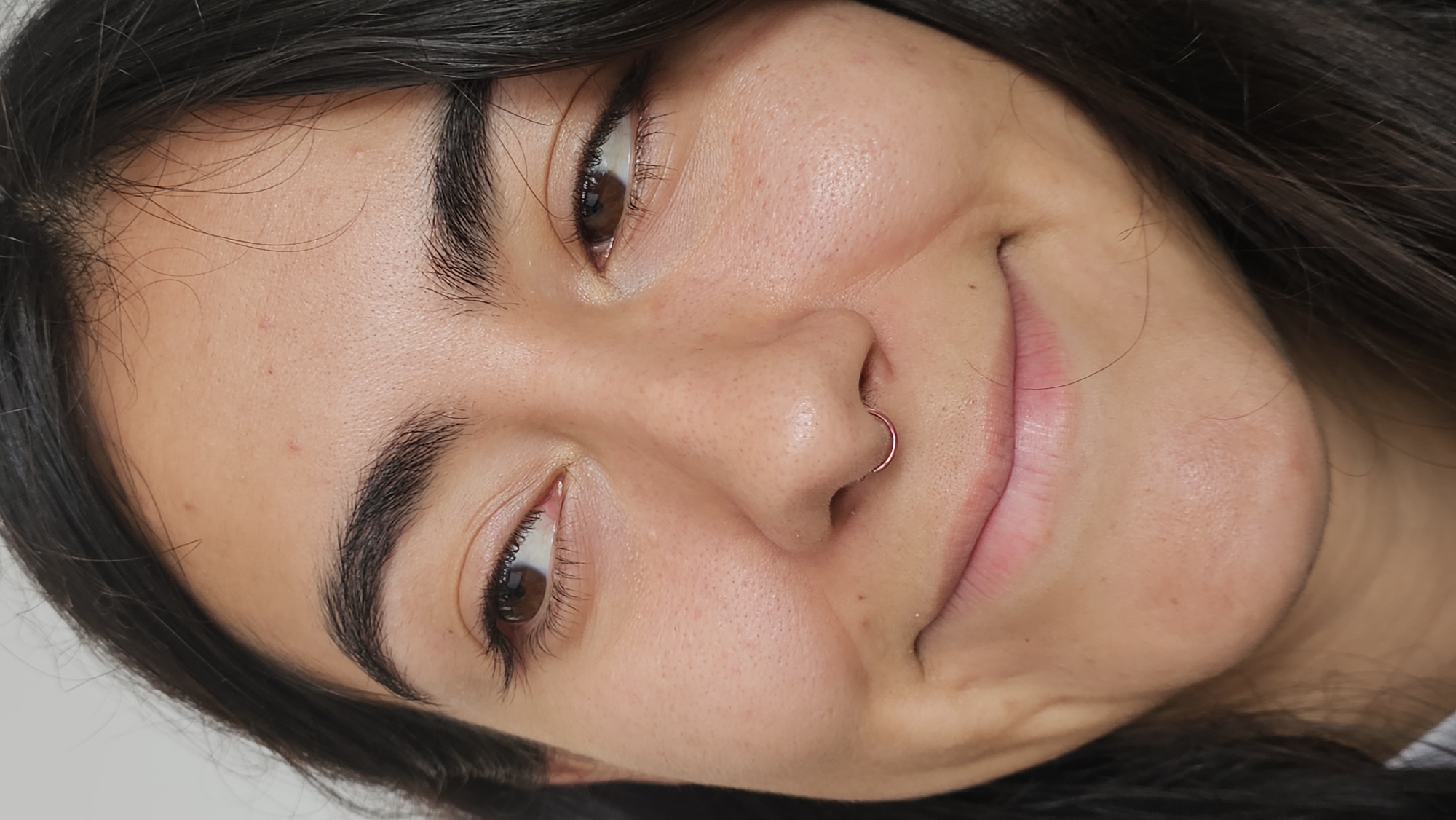 A close-up shot of a woman with brown eyes, dark hair, and a nose ring. The woman is smiling slightly, and the image is taken from a low angle. The background is out of focus. The location is not discernible from the image.