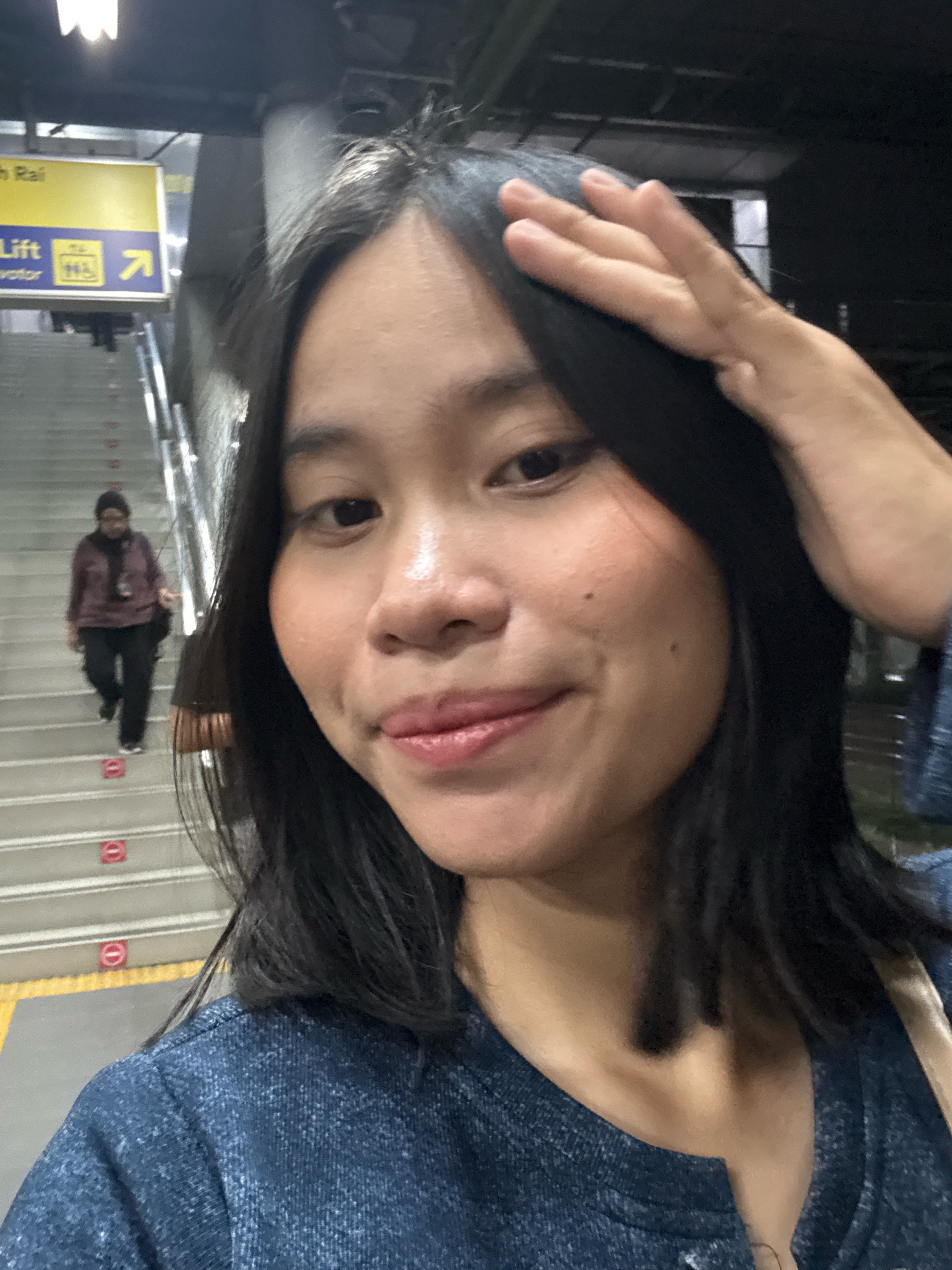 A selfie is taken of a young woman with dark hair, reaching up to touch her hair. She is wearing a blue denim jacket. Behind her, a woman in a maroon jacket and black pants walks down a staircase. A yellow sign with "Lift" and an arrow is visible. The scene appears to be a public area with stairs and signage, possibly a transit station in East Jakarta, Indonesia.