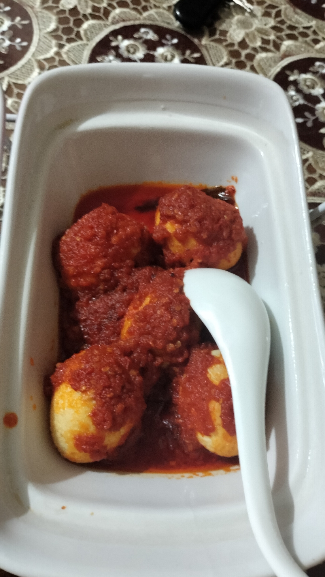 A white rectangular dish containing boiled eggs in a spicy red sauce is placed on a tablecloth. A white spoon rests on the dish. The image was taken in a home in Lhokseumawe, Indonesia.