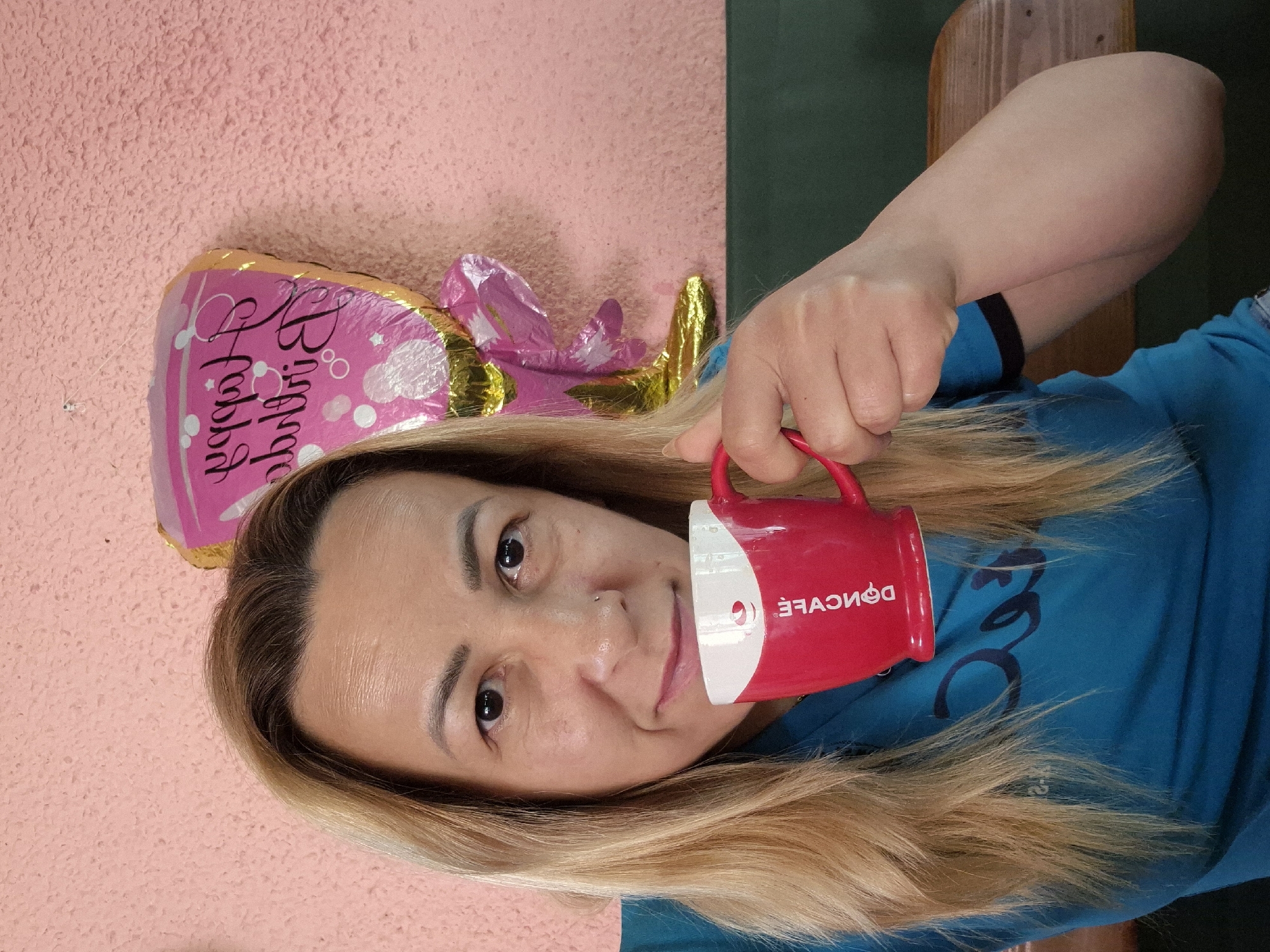 A woman with blonde hair and a blue shirt holds a red and white mug to her face, taking a drink. Behind her, a pink wall with a textured surface is visible, along with a pink and gold balloon that says "Happy Birthday". The lower portion of the image shows a dark green surface and a wooden bench.