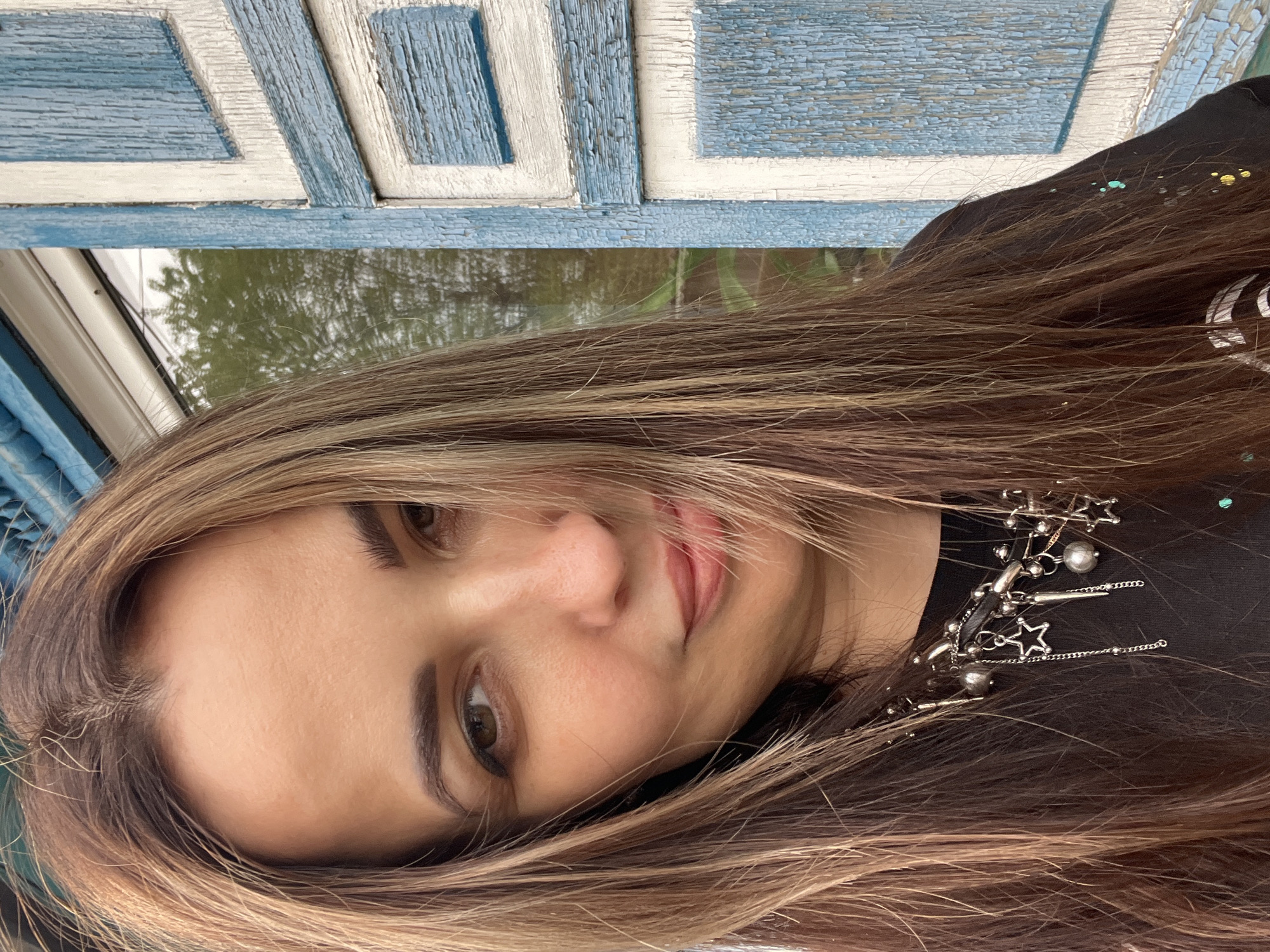 A woman with shoulder-length brown hair and light blonde highlights, wearing a black top and a silver star and pearl necklace, is positioned in front of a weathered blue and white patterned wooden structure, possibly a window or door frame. Behind the structure is greenery, suggesting an outdoor location, possibly in Unknown, Unknown. The image is slightly tilted.