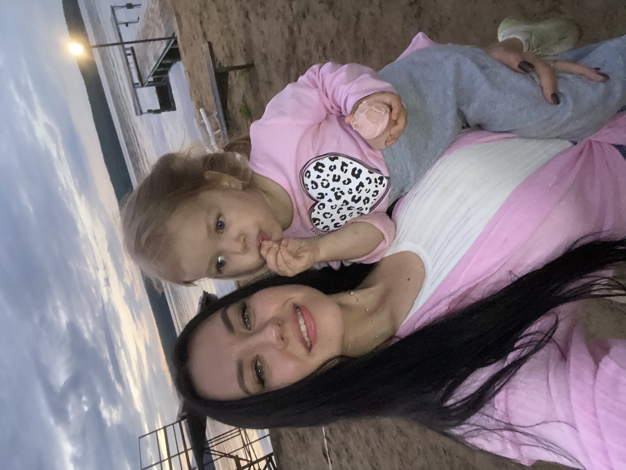 In a selfie taken outdoors, likely near the Kama River in Chaikovsky, Russia, a woman with long, dark hair smiles at the camera, holding a young child. The child, with blonde hair, is being held in the woman's arms and appears to be eating something. The child is wearing a pink long-sleeved shirt with a leopard-print heart design and grey sweatpants, and is holding a pink scoop of food. The woman is wearing a white top and a pink cardigan or shawl. In the background, a body of water, possibly the Kama River, is visible under a cloudy sky. A wooden pier and a lamppost are also visible on the shore, suggesting a recreational area. The ground is sandy, typical of a riverbank or beach.