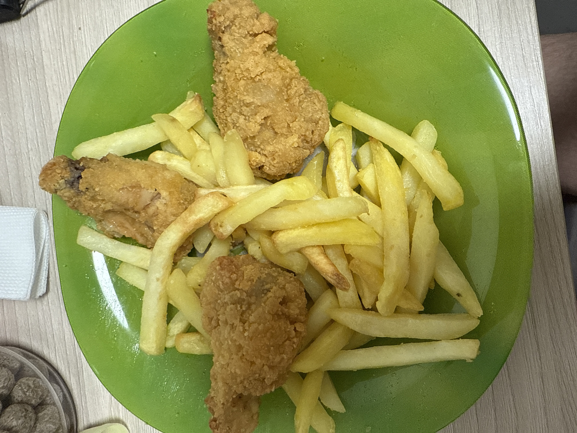 A bright green plate is filled with french fries and fried chicken. Three pieces of fried chicken are visible, one drumstick at the top of the plate and two pieces that appear to be wings. The french fries are golden brown and fill the majority of the plate. The food is laid out on a light wood-grain surface. To the left of the plate, a white paper napkin is folded. Below the napkin and to the left of the plate, a clear plastic container holds small, round, brown objects that appear to be food. The lighting suggests it is daytime, possibly indoors, as there are no visible windows or outdoor elements. The scene is a close-up shot of the food, with no people visible. The image is taken from directly above the plate.