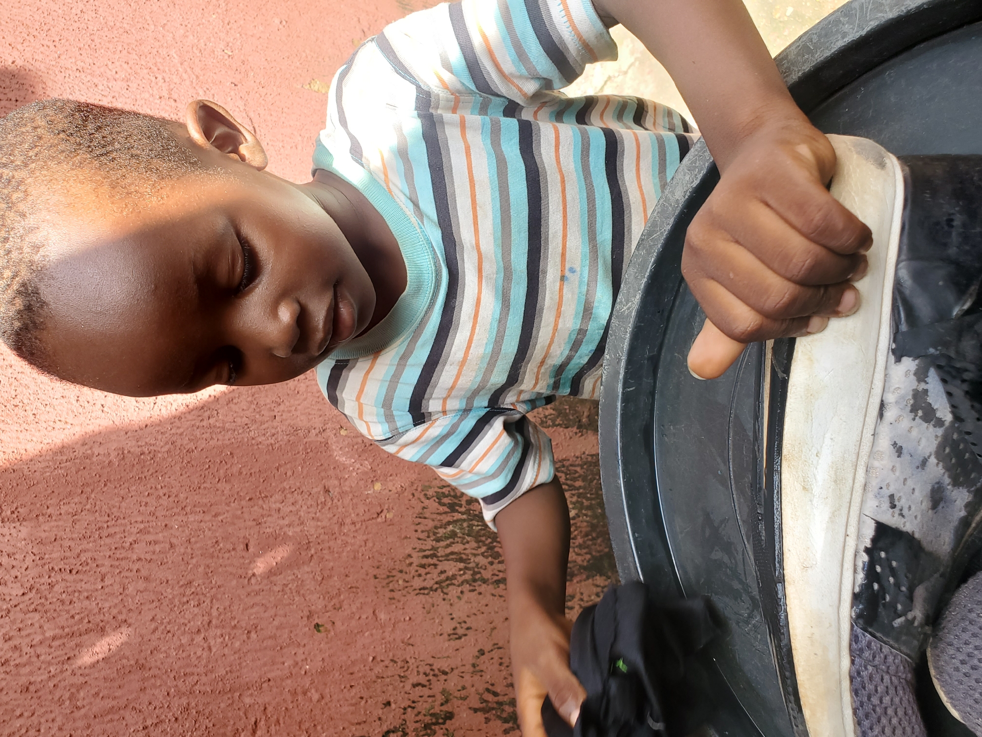 The image shows a young Black child, likely a boy, from a high angle. He is wearing a striped t-shirt with horizontal bands of light blue, orange, dark blue, and grey. The child has his eyes closed and appears to be concentrating. Sunlight casts a distinct shadow across his face and hair. He is reaching into a large black plastic container, possibly a bucket or a wash basin. In his left hand, he is holding a black piece of fabric. His right hand is resting on the rim of the container, near a dirty white sole of a shoe or similar object. The background is a textured, reddish-brown surface, likely the ground, with some small patches of green, possibly moss or weeds. The overall lighting suggests it is daytime, with the sun high in the sky. The scene evokes a sense of focused activity, possibly washing or cleaning an object.