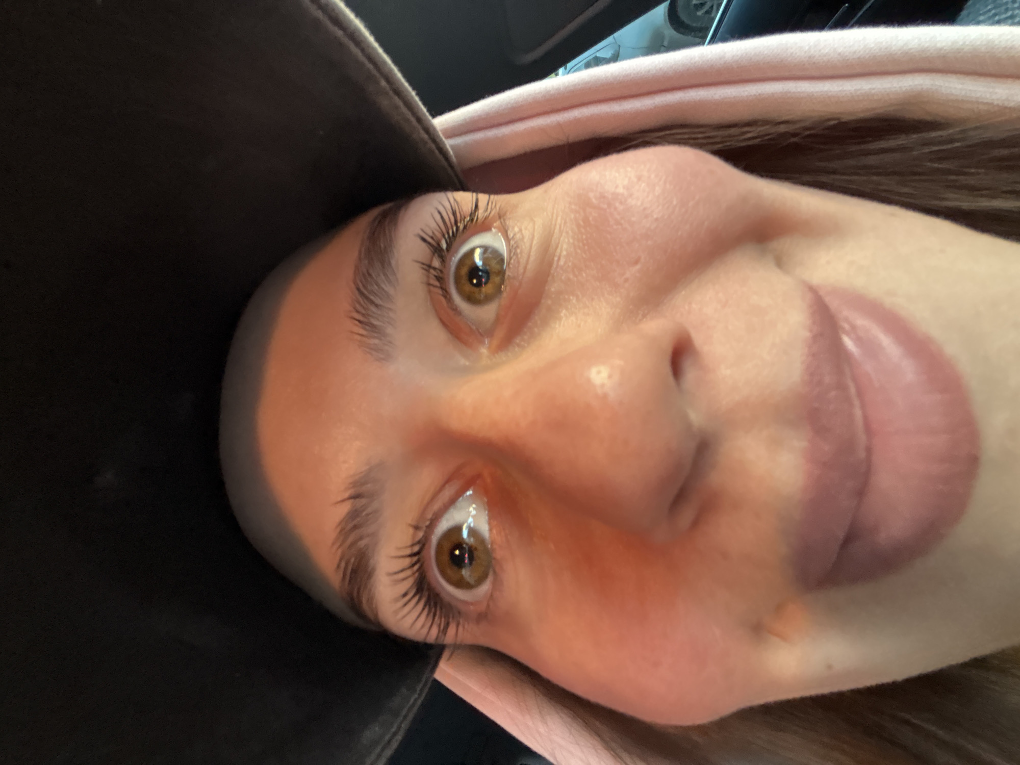 A close-up, upward-angled selfie shows a fair-skinned person wearing a black cap and a light pink hooded garment. The individual has brown eyes, prominent eyelashes, and full lips that are pursed. The background is partially visible and suggests the inside of a car. There is no indication of the location being Krasnoyarsk, Russia.