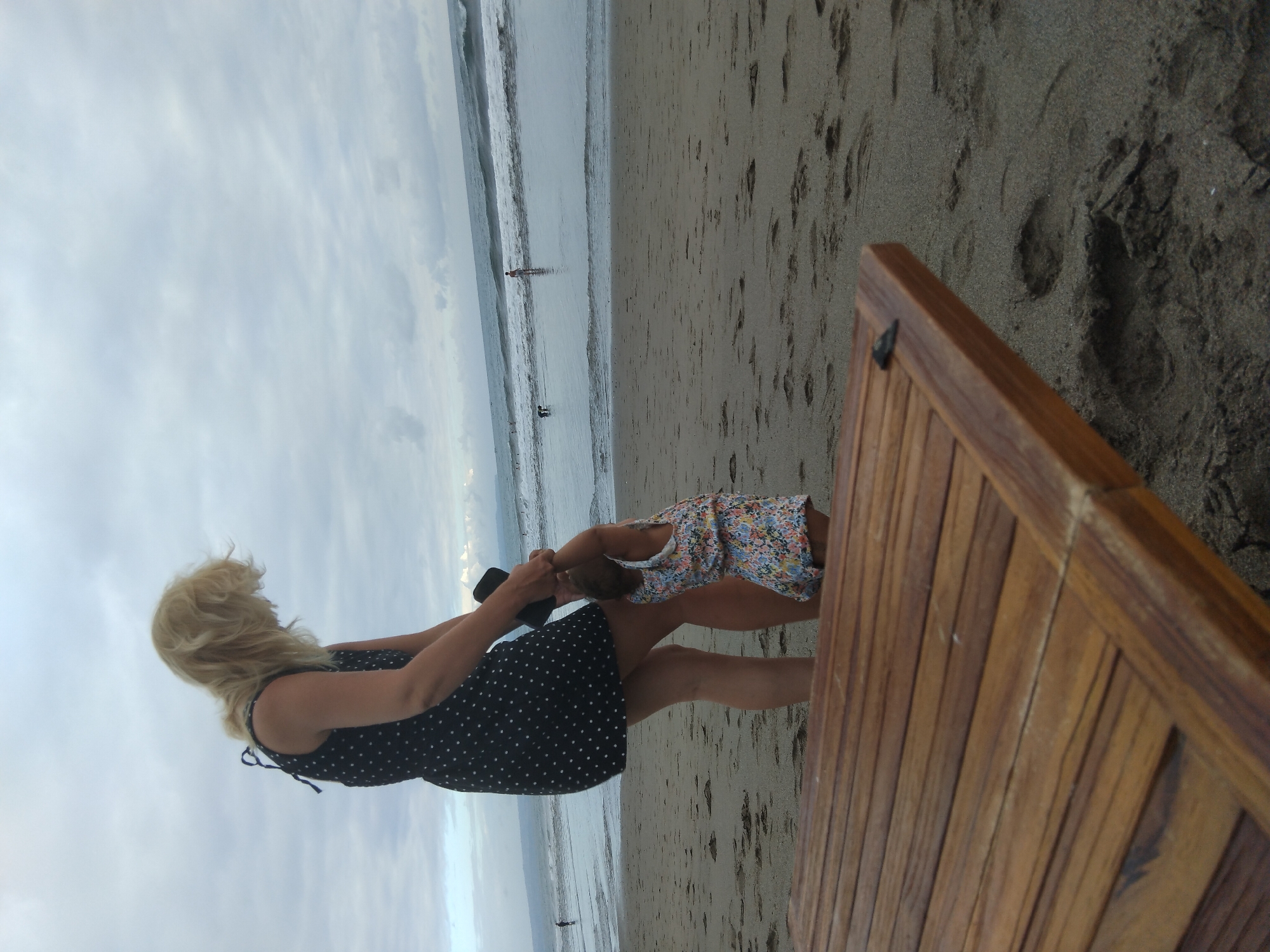 The image shows two people on a beach in North Kuta, Indonesia. A woman with blonde hair, wearing a black polka dot dress, is holding the hand of a young child. The child is wearing a colorful floral dress. They appear to be playing or interacting on the sandy beach. In the background, the ocean is visible with waves, and there are a few other people in the water. The sky is overcast, suggesting a cloudy day, possibly early morning or late afternoon. A wooden structure, possibly a bench or table, is prominently in the foreground, partially obscuring the view. The scene depicts a moment of leisure and family time on a tropical beach.