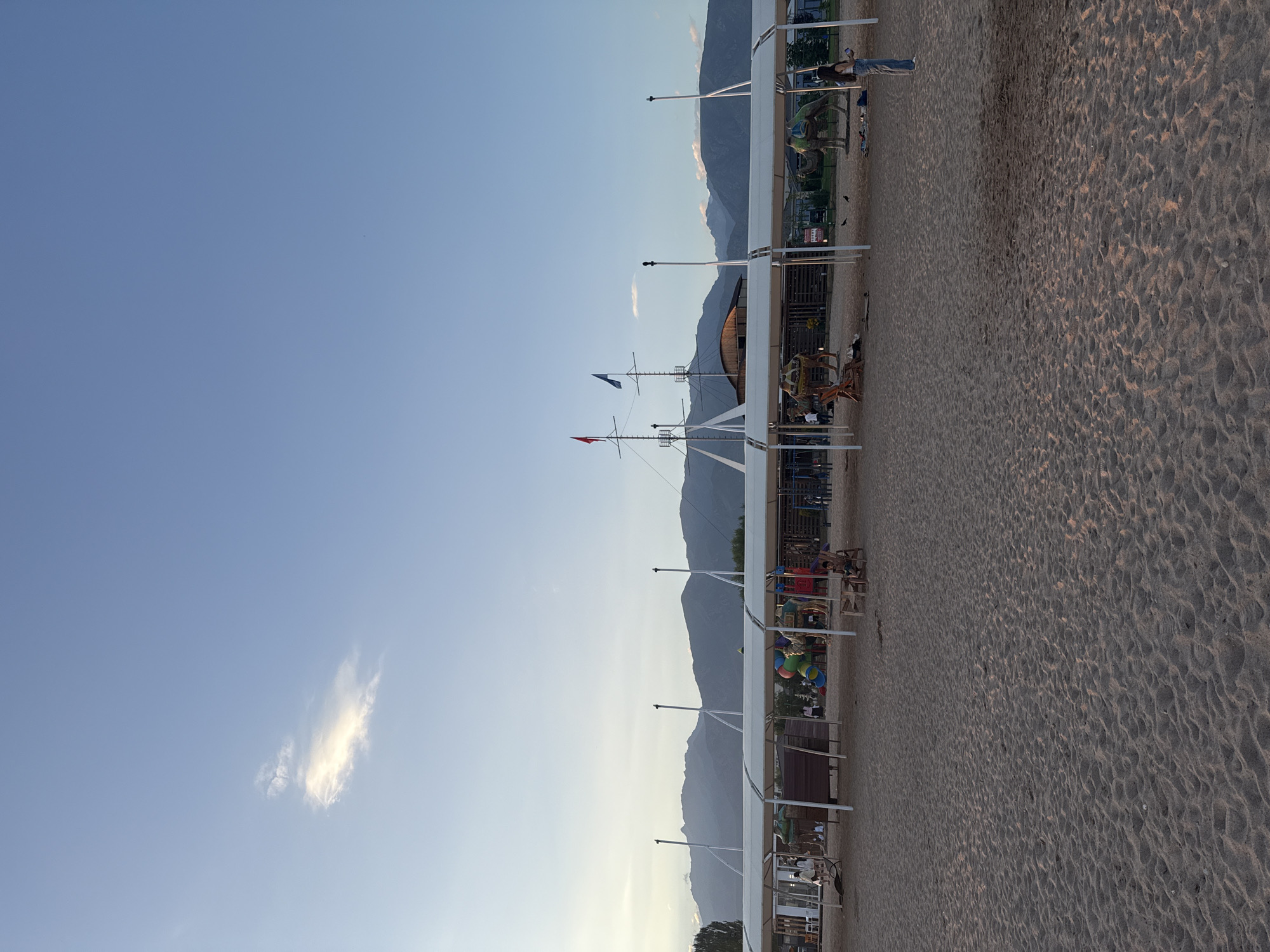 The image depicts a sandy beach area with a clear blue sky and a distant mountain range. In the foreground, the textured sand is illuminated by soft, natural light, suggesting it is either early morning or late afternoon. A white canopy structure stretches across the middle ground, providing shade for a playground area with colorful slides and climbing structures, and what appear to be camel statues or props, one adorned with a saddle. Several people are scattered throughout the scene, some sitting on benches under the canopy, others walking or standing on the sand. In the background, a large, ship-like structure with masts and flags is visible, adding a unique element to the beach setting. The overall atmosphere is relaxed and recreational, characteristic of a resort or park area. The location is identified as the city of Sary-Oy, Kyrgyzstan, a known resort town on the shore of Lake Issyk-Kul.