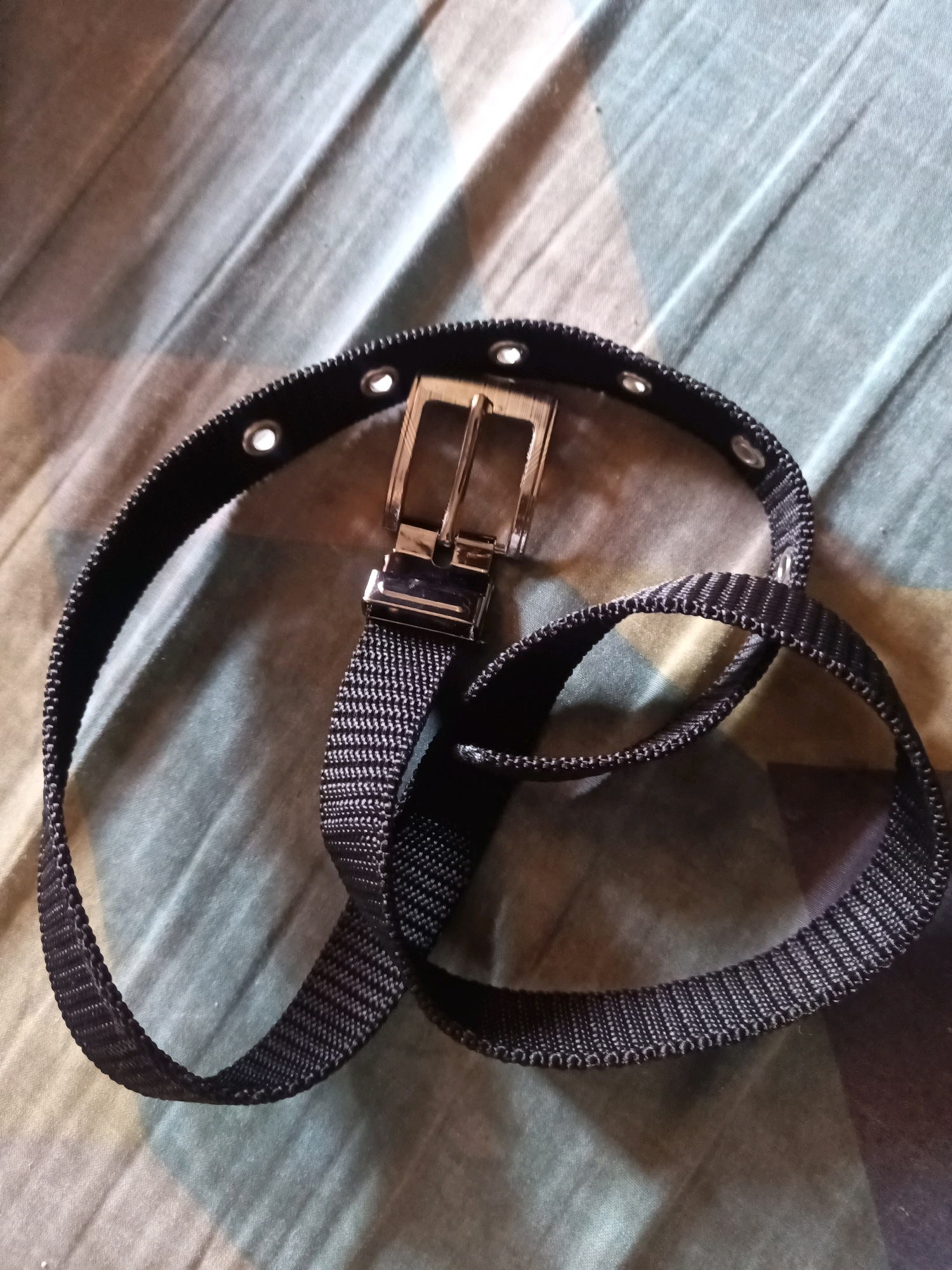 A black belt with a silver buckle and eyelets is draped over a fabric surface. The fabric has a diagonal striped pattern in shades of green and cream. The belt is coiled and appears to be made of a woven material.