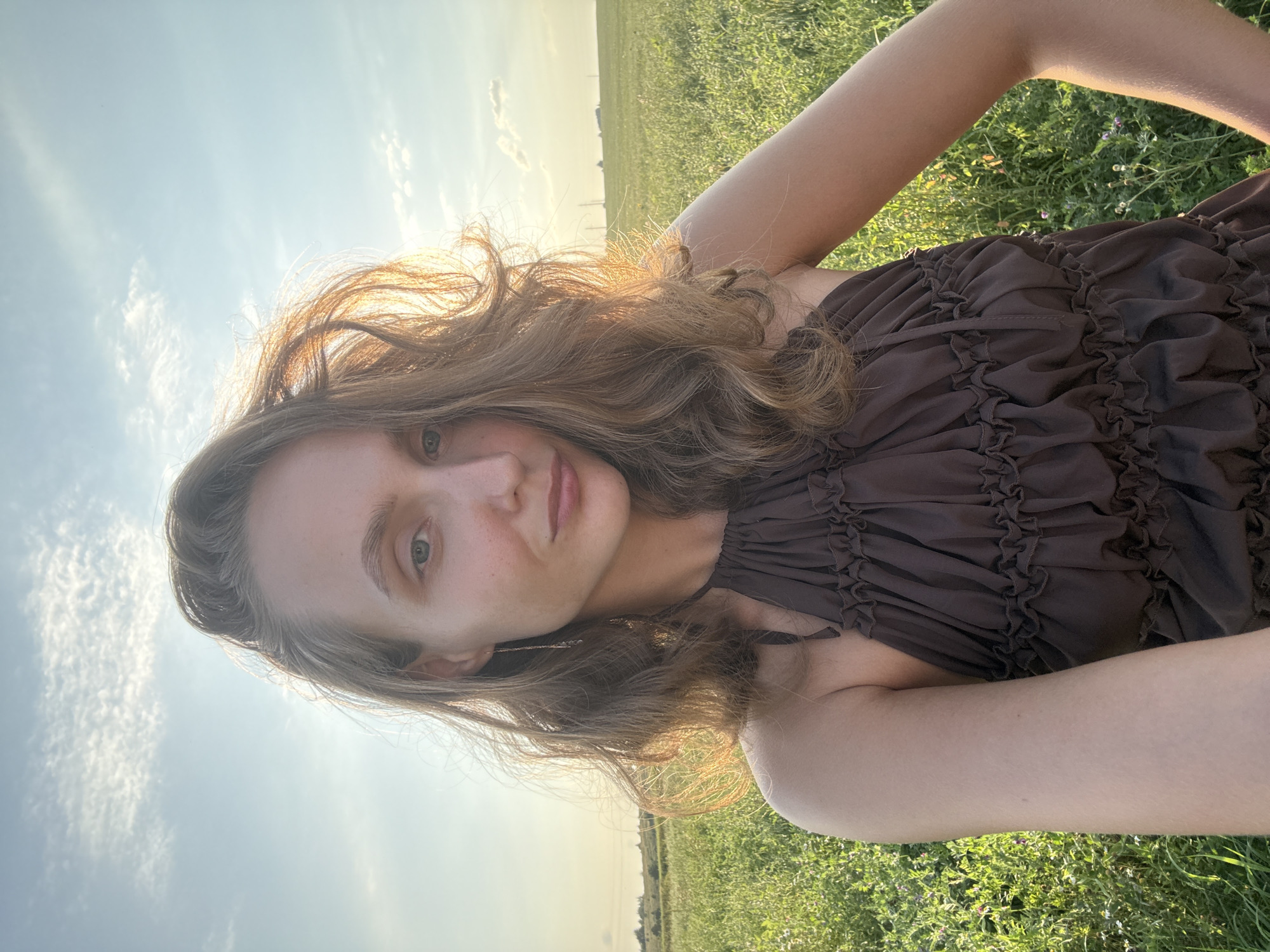 A young woman is taking a selfie in a sunlit field. She has long, wavy brown hair and is wearing a brown, ruffled halter dress. Her left arm is bent, with her hand resting on her hip. The background shows a vast, green field under a bright sky with scattered clouds. The scene appears to be in Aksaitovo, Russia, given the landscape.