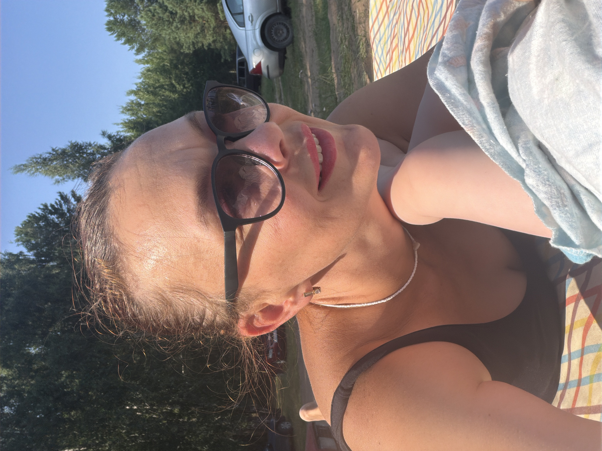 The image shows a woman lying down outdoors, with trees and a clear blue sky in the background. She is wearing a black tank top, sunglasses, and a white beaded necklace with a small pendant. Her hair is pulled back, and she is resting her head on her arm. A blue towel is draped over her legs, and she is lying on a striped blanket. The scene appears to be a park or a recreational area. The location is described as Noviy, Russia.