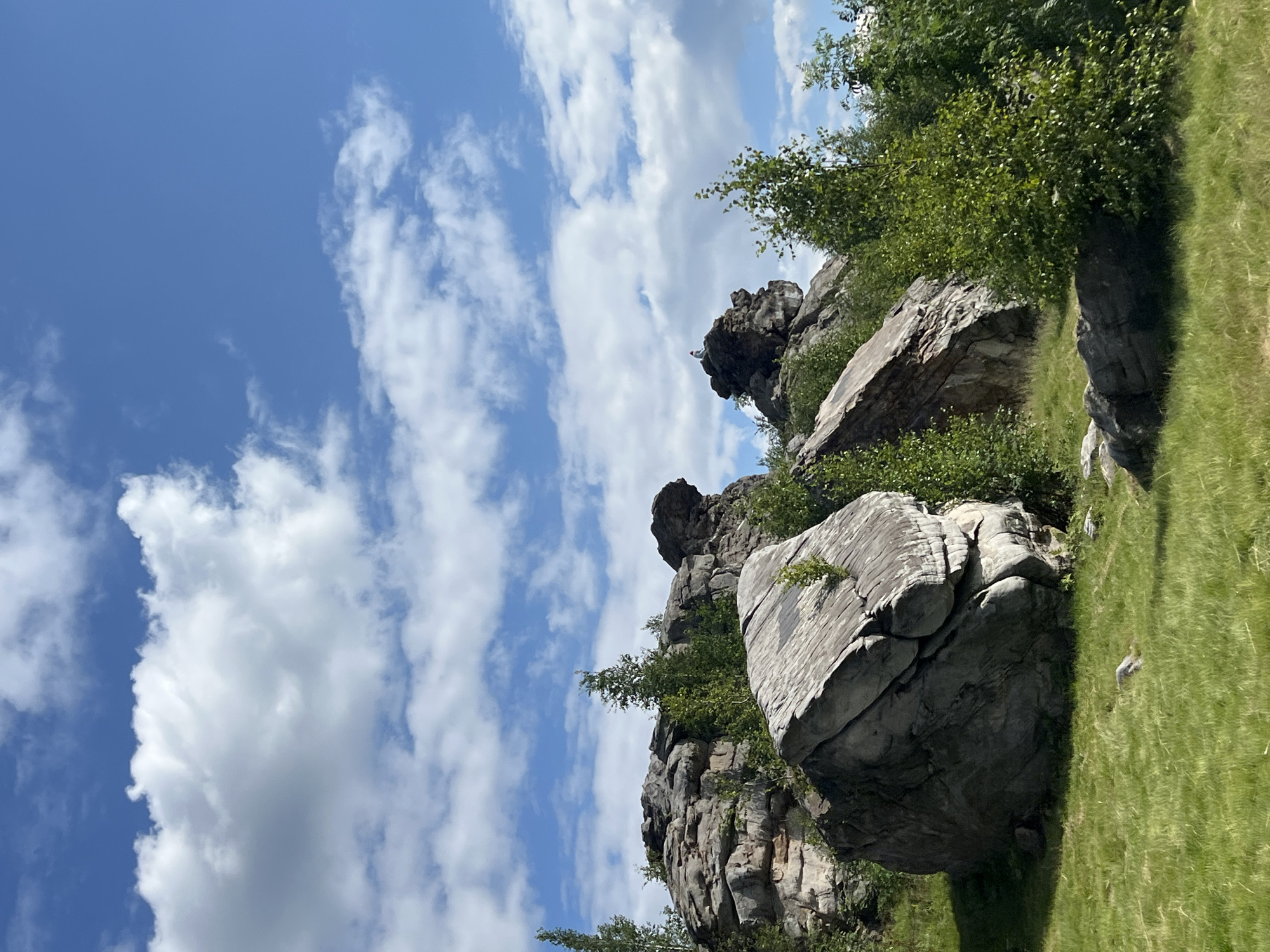 The image shows a landscape with large rock formations and lush green vegetation under a bright blue sky with scattered white clouds. In the foreground, a large, weathered boulder with visible striations lies on a grassy slope. Behind it, more massive rock formations rise, interspersed with trees and bushes. The rocks are gray and appear to be composed of layered sedimentary material. To the right, a person is visible sitting atop one of the rock formations, appearing small in relation to the landscape. The scene is likely located at the "Forbidden Forest" (Заповедный лес) rock formations near Gubakha, Russia, which are known for their unique geological structures and scenic beauty.