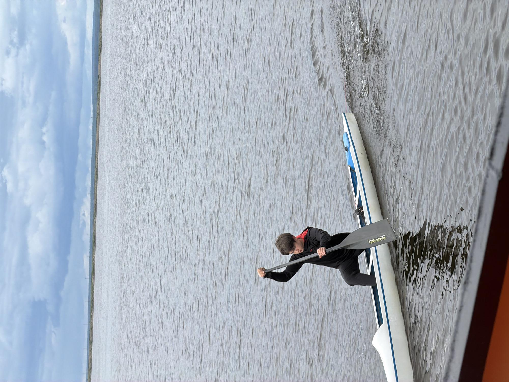 A man is rowing a boat on the water. The boat is white with a blue stripe, and he is holding a paddle. The water is grey and choppy, and there is a cloudy sky in the background. The scene appears to be taking place in the city of Chaikovsky, Russia.