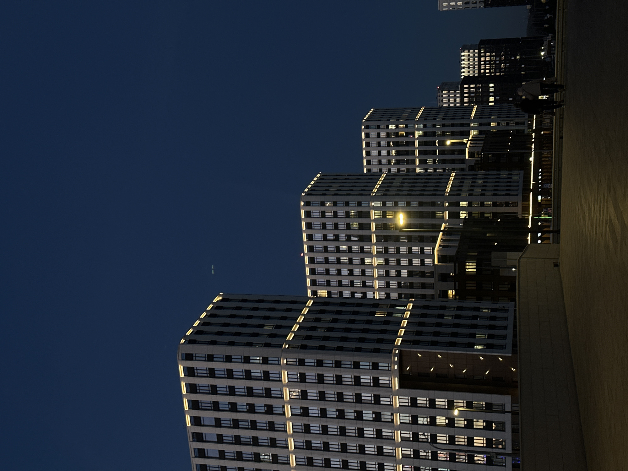 The image presents a modern urban landscape at night in Kazan, Russia, dominated by several brightly illuminated high-rise buildings against a deep blue, clear sky. The buildings, featuring contemporary architecture, rise in a staggered, receding formation from left to right across the frame.
These multi-story structures display numerous rectangular windows, many of which glow with warm, artificial light from within. A striking detail is the extensive exterior strip lighting that outlines the various tiers and edges of the buildings in bright, warm white, creating a strong geometric pattern and enhancing their modern aesthetic. A particularly intense light source, likely a streetlamp, is visible nestled between two of the buildings.
On the right side of the image, a wide, paved area, possibly a sidewalk or promenade, stretches horizontally. It is brightly illuminated by ambient light, giving it a warm, yellowish tone. Dark silhouettes of a few people are visible walking along this path, providing a sense of scale and human activity within the urban environment. The sky is uniformly dark blue, signifying nighttime, with no clouds visible, though a faint, thin light streak is discernible in the upper left, possibly from an aircraft. The scene conveys the vibrant and active atmosphere of a city after dark.
