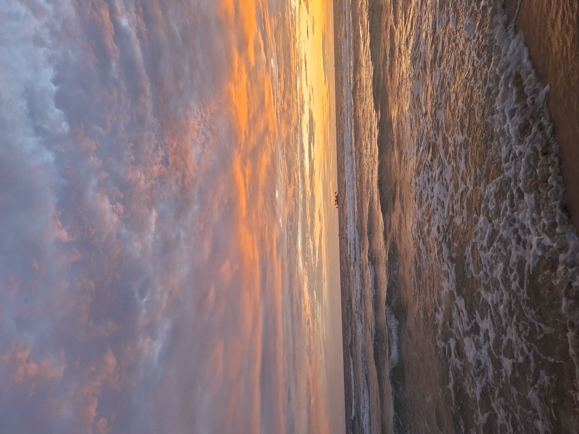 This image depicts a sunset over the ocean, likely on the western coast of Phú Quốc, Vietnam, where such views are common. The sky is a vibrant canvas of orange, pink, and purple hues, with scattered clouds reflecting the warm light of the setting sun. Gentle waves roll towards the sandy shore, creating white foam as they break. In the distance, a few small boats can be seen on the horizon. The overall scene is one of natural beauty and tranquility, characteristic of a tropical sunset.