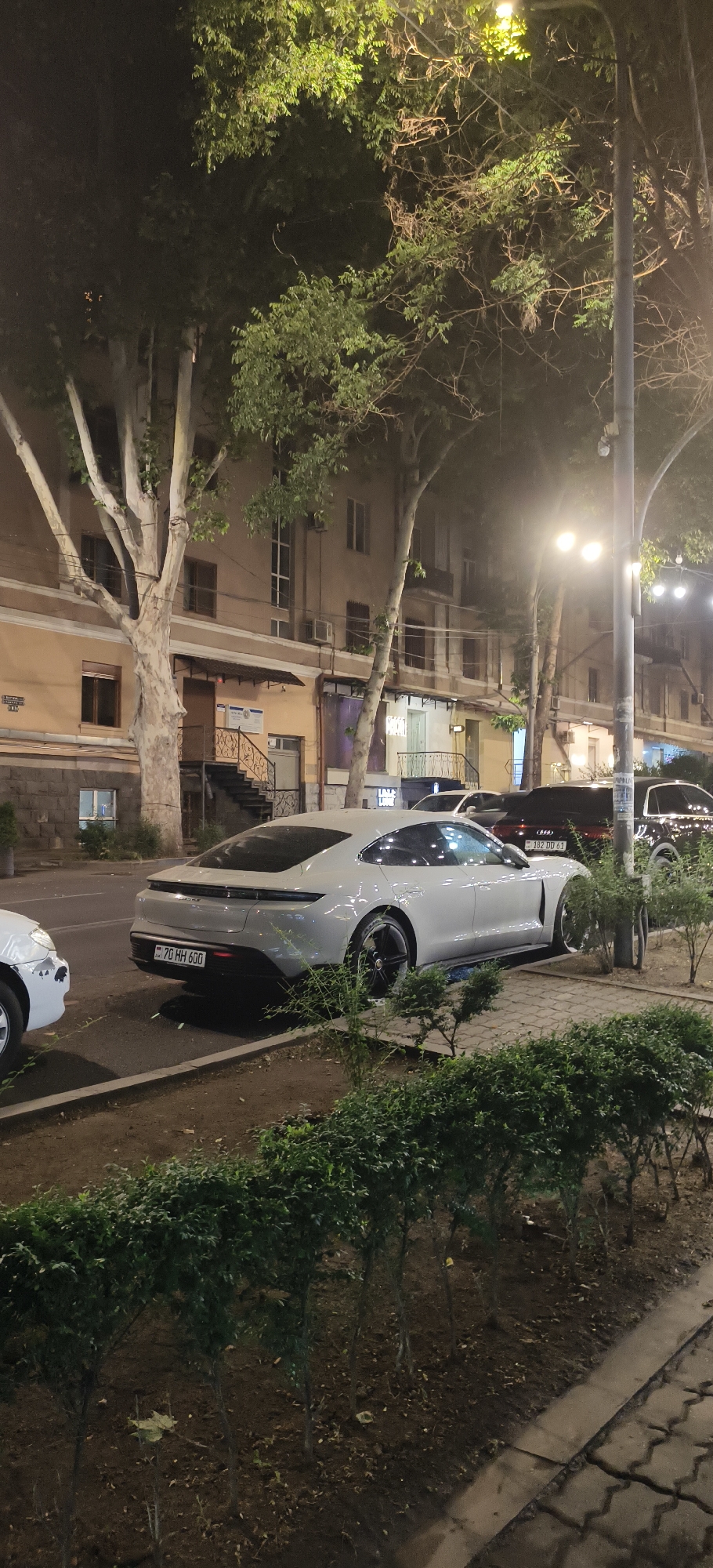 The image depicts a street scene at night, with a white electric car, identified by its license plate "70 HH 600", parked on the side of the road. The car is a Porsche Taycan, and it is facing left. Next to it, to the right, are other vehicles, including a dark-colored SUV with license plate "382 DD 61". In the background, there is a multi-story building with several windows and balconies, and trees line the sidewalk, their leaves illuminated by streetlights. The scene suggests an urban environment at night, possibly a residential or commercial area.