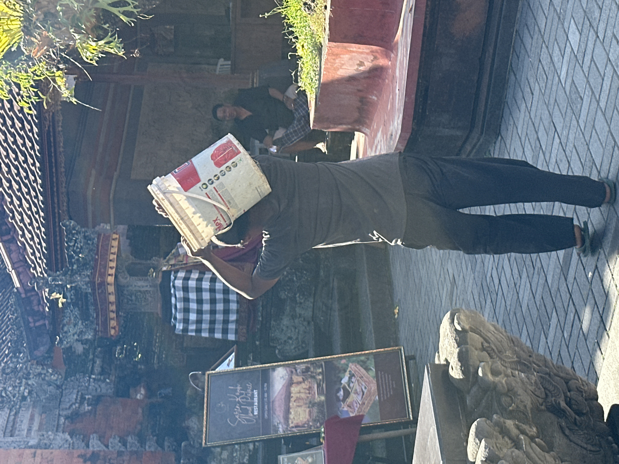 In Ubud District, Indonesia, a man is seen carrying a white bucket with red markings on his head. He is wearing a dark grey t-shirt and dark pants. The man is walking on a paved area with rectangular grey tiles. To the left, a tall framed sign displays text and images, including a picture of a building and a basket of food. Behind the man, a checkered black and white cloth is visible, draped over something. Further in the background, another man is seated, wearing a dark shirt and plaid pants. The environment appears to be an outdoor setting with architectural elements suggesting a traditional Balinese style, including a tiled roof and ornate stonework. Green plants are also present in the scene. The lighting suggests it is daytime.