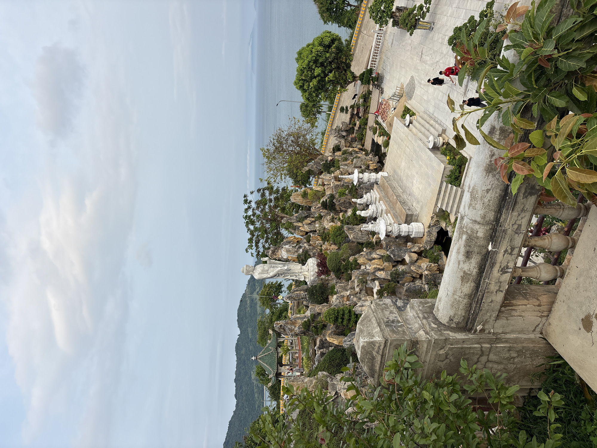 The image shows a view from above, looking down at a landscaped area with a large white statue, likely the Lady Buddha statue, within the Linh Ung Pagoda complex on the Sơn Trà Peninsula in Đà Nẵng, Vietnam. The ocean and a cloudy sky are in the background. Several stone columns and structures are visible. A group of people is walking in the upper right part of the image. Green foliage and vegetation fill the foreground and parts of the midground. There are a few pavilions throughout the complex.