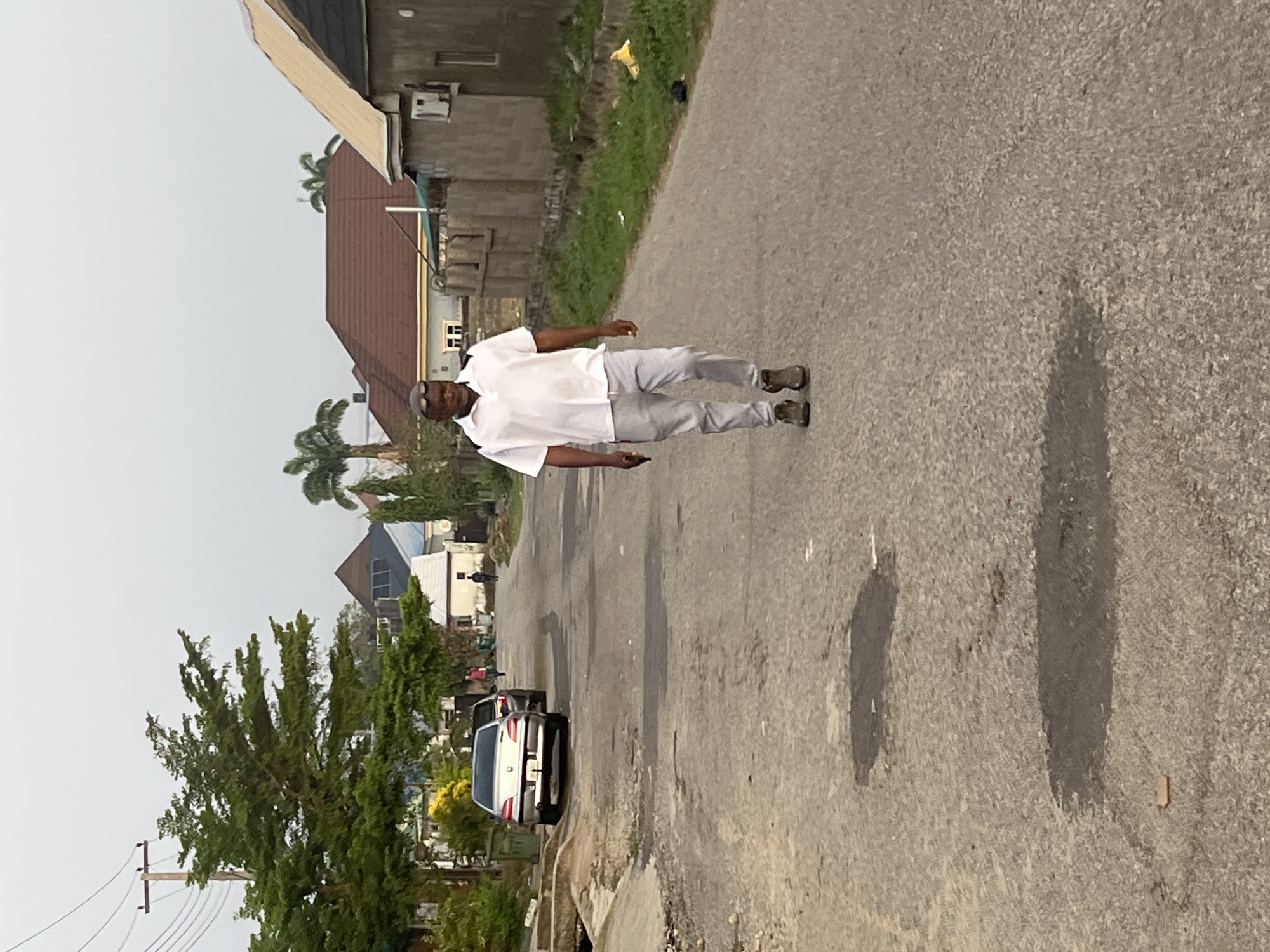 A man is walking down a paved street in an urban area. He is wearing a light-colored short-sleeved shirt, light-colored pants, and dark shoes. He appears to be looking down. A car is parked on the side of the street behind him. There are buildings and trees visible in the background. The scene is in Abuja, Nigeria.