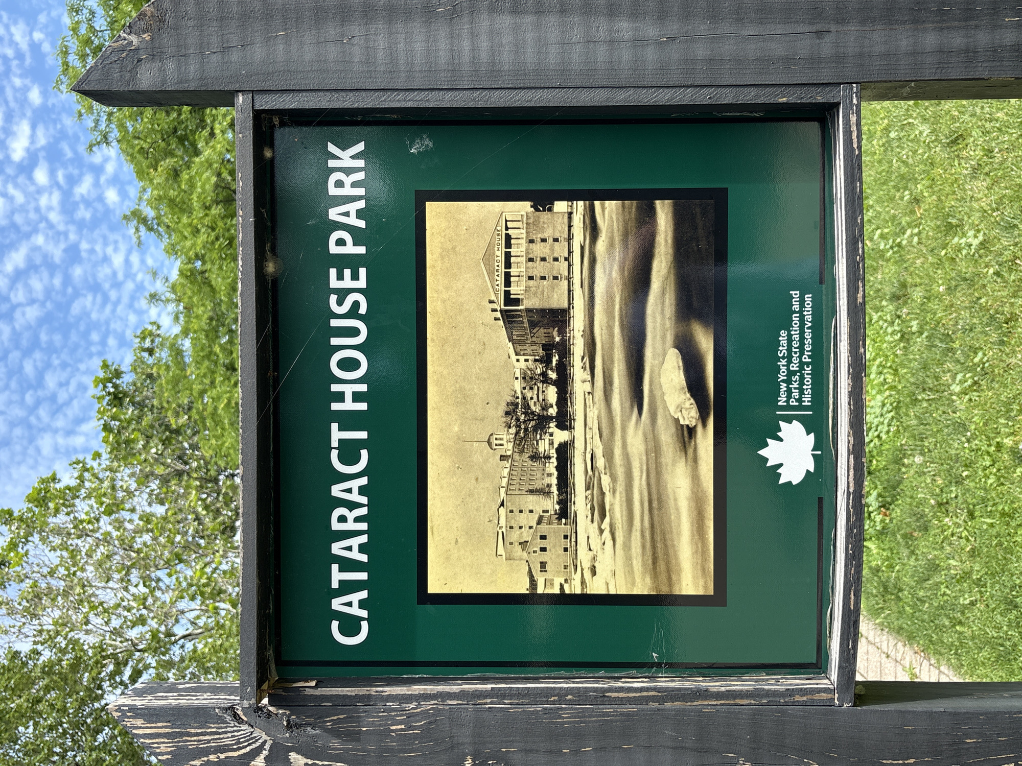 A green sign with white lettering reads "CATARACT HOUSE PARK" at the top. Below the text is a historical sepia-toned photograph of a large building complex with a dome and the words "CATARACT HOUSE" visible on one of the structures. The photograph depicts a body of water or icy landscape in the foreground. At the bottom of the sign, a white maple leaf logo is to the left of text that reads "New York State Parks, Recreation and Historic Preservation". The sign is mounted on a weathered wooden post. The background shows green trees and a partly cloudy blue sky. The sign is located in the City of Niagara Falls, United States of America.