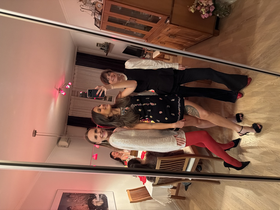 A mirror reflects three women in evening wear. The first, wearing a sparkly top and red tights, sits on a chair. Above her, a woman in a black dress with floral details stands, showcasing a leg tattoo. The third woman, in a white blouse and black suit, holds a phone, taking a selfie. The reflection also reveals a room interior, including a wooden dresser, artwork, lighting fixtures, and decorative flowers.