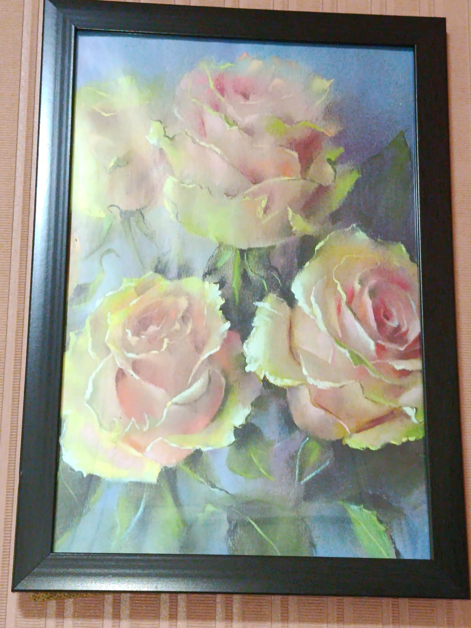 The image displays a framed painting of three light pink and yellow roses against a dark blue and green background. The painting is hanging on a wall with a textured, light brown wallpaper. The frame is dark brown with a visible wood grain. No specific location in Tashkent, Uzbekistan is identifiable.