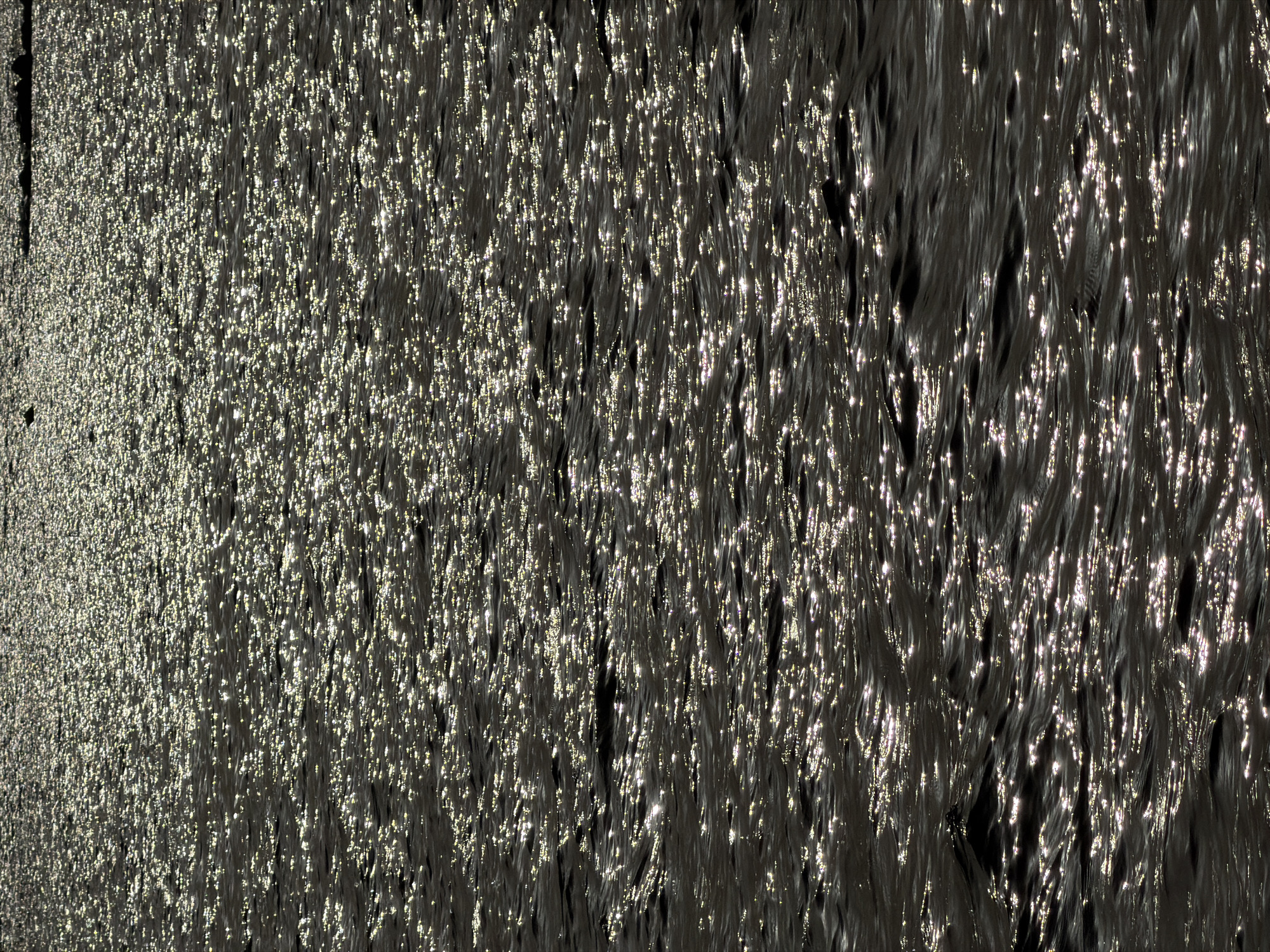 This image captures the sunlit surface of a body of water, likely the sea or a large lake. The water is dark and choppy, with a multitude of small, bright reflections scattered across it, indicating a sunny day with a light breeze. The reflections create a shimmering, textured effect. In the distance, along the horizon, a dark, indistinct line suggests land or possibly a rocky shoreline. The overall impression is one of natural beauty and the play of light on water. This scene is likely to be found along the coast of Alghero, Italy, a city known for its beautiful Mediterranean coastline. There are no people or specific man-made objects visible in this particular frame.
