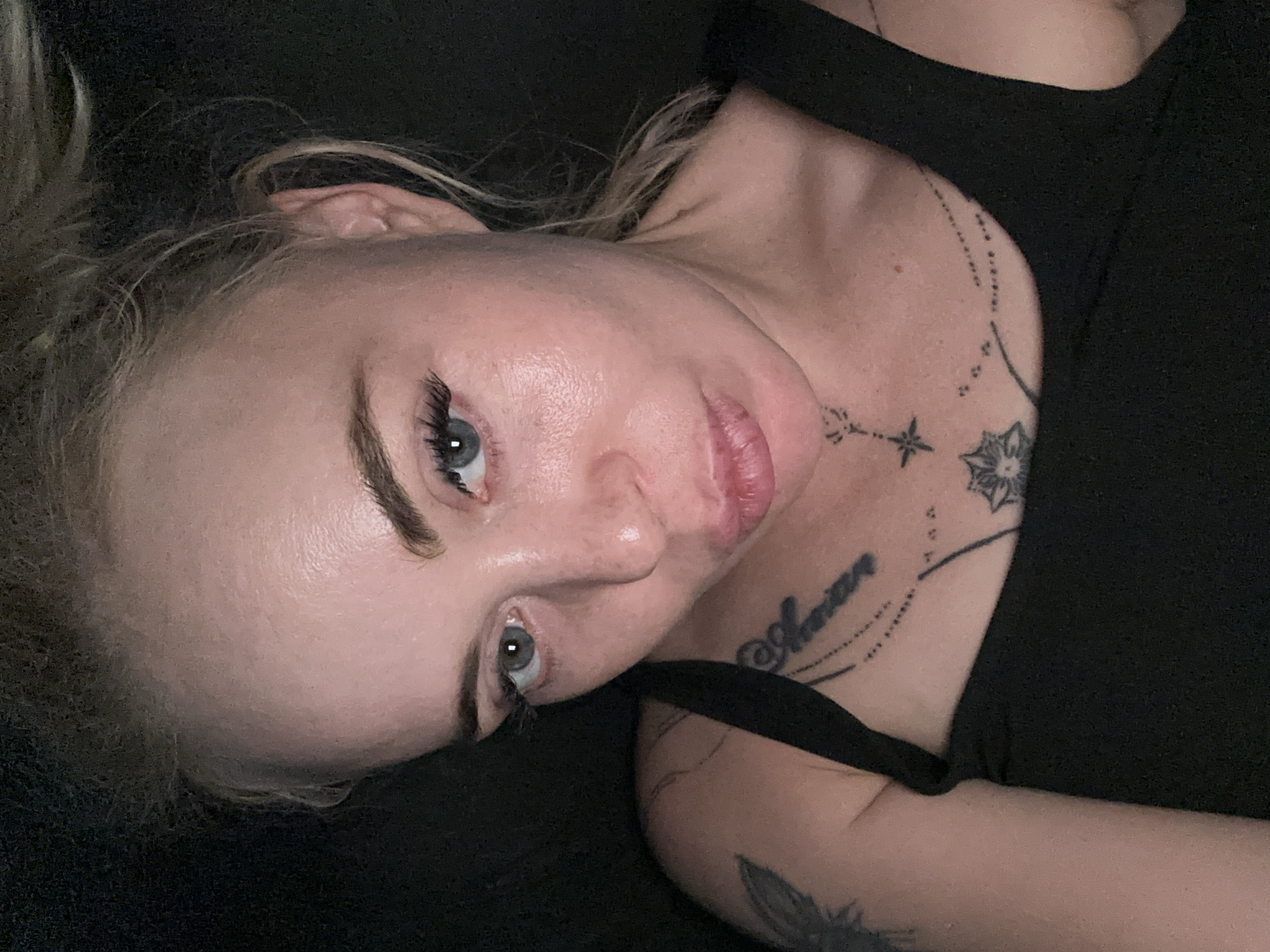 A close-up, overhead shot shows a woman with blonde hair pulled back from her face, looking down at the camera. She has light blue eyes with dark eyelashes, prominent eyebrows, and fair skin with a slight sheen. Her lips are slightly parted and appear to be painted with a light pink lipstick. She is wearing a black tank top, and tattoos are visible on her chest and left arm. The background is dark and out of focus, suggesting an indoor setting with low lighting.