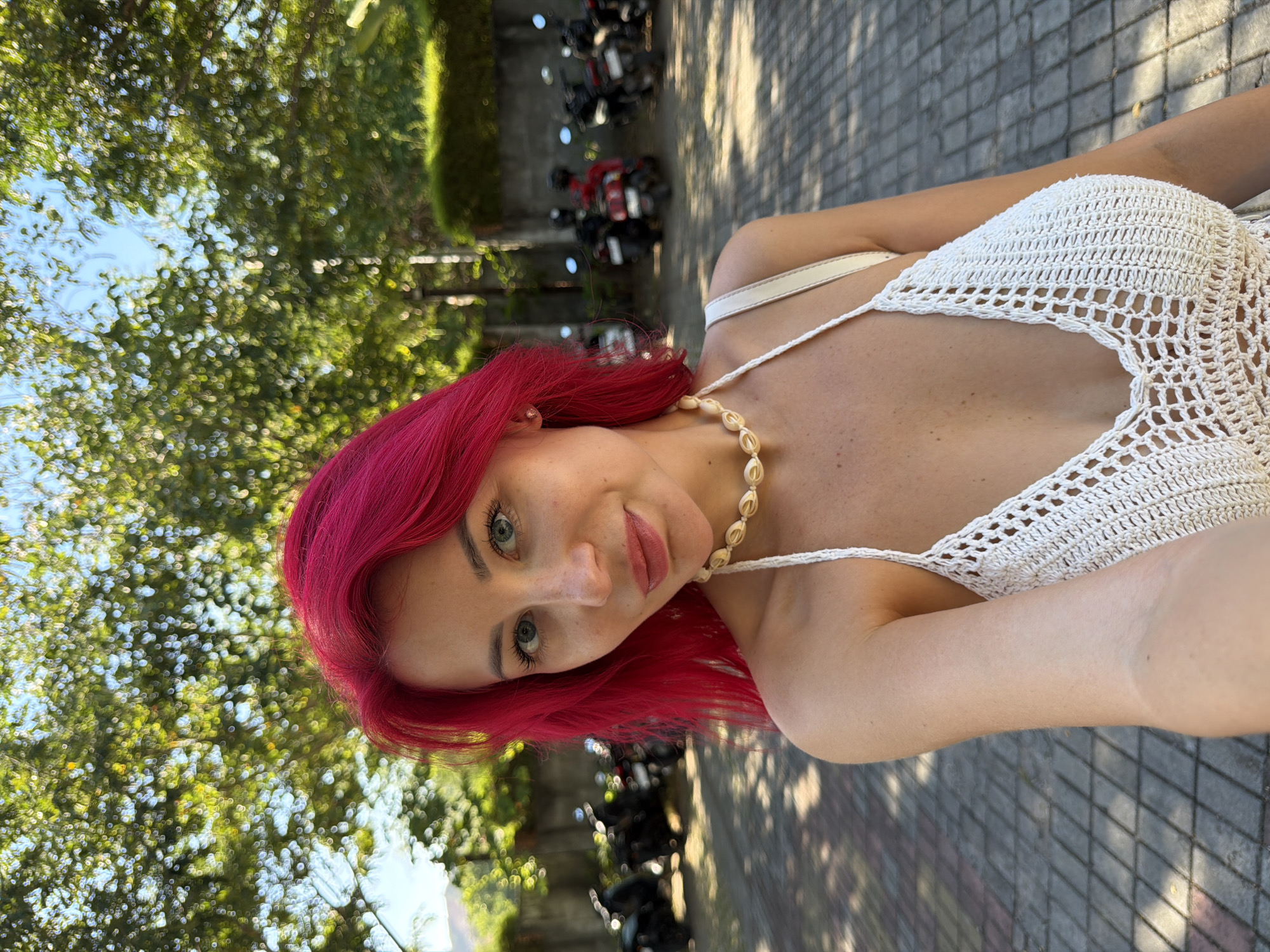 A young woman with bright magenta hair and blue eyes is captured in a selfie in Kuta, Indonesia. She is wearing a white crocheted top and a seashell necklace. The background shows a paved area with parked motorcycles and lush green trees. The lighting suggests it is daytime and sunny.