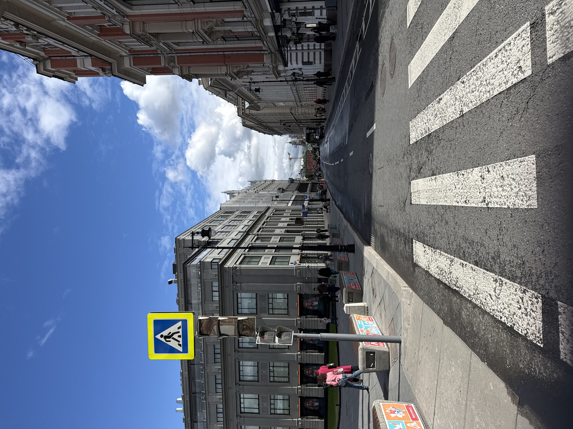 This photograph depicts a street scene in Moscow, Russia, notable for its architecture, pedestrian activity, and urban infrastructure.
The foreground features a pedestrian crosswalk with white stripes on a gray asphalt road. A blue and yellow pedestrian crossing sign is visible. People are present on the sidewalk, including a woman in a pink shirt and jeans.
Buildings line both sides of the street, showcasing different architectural styles. The building on the left side is predominantly gray with numerous windows, while the buildings on the right exhibit a more ornate design with red and white facades.
The sky is blue with scattered white clouds, providing a bright backdrop to the urban environment. In the distance, the street extends into the horizon, with additional buildings and pedestrian activity visible. The overall scene suggests a typical day in Moscow.