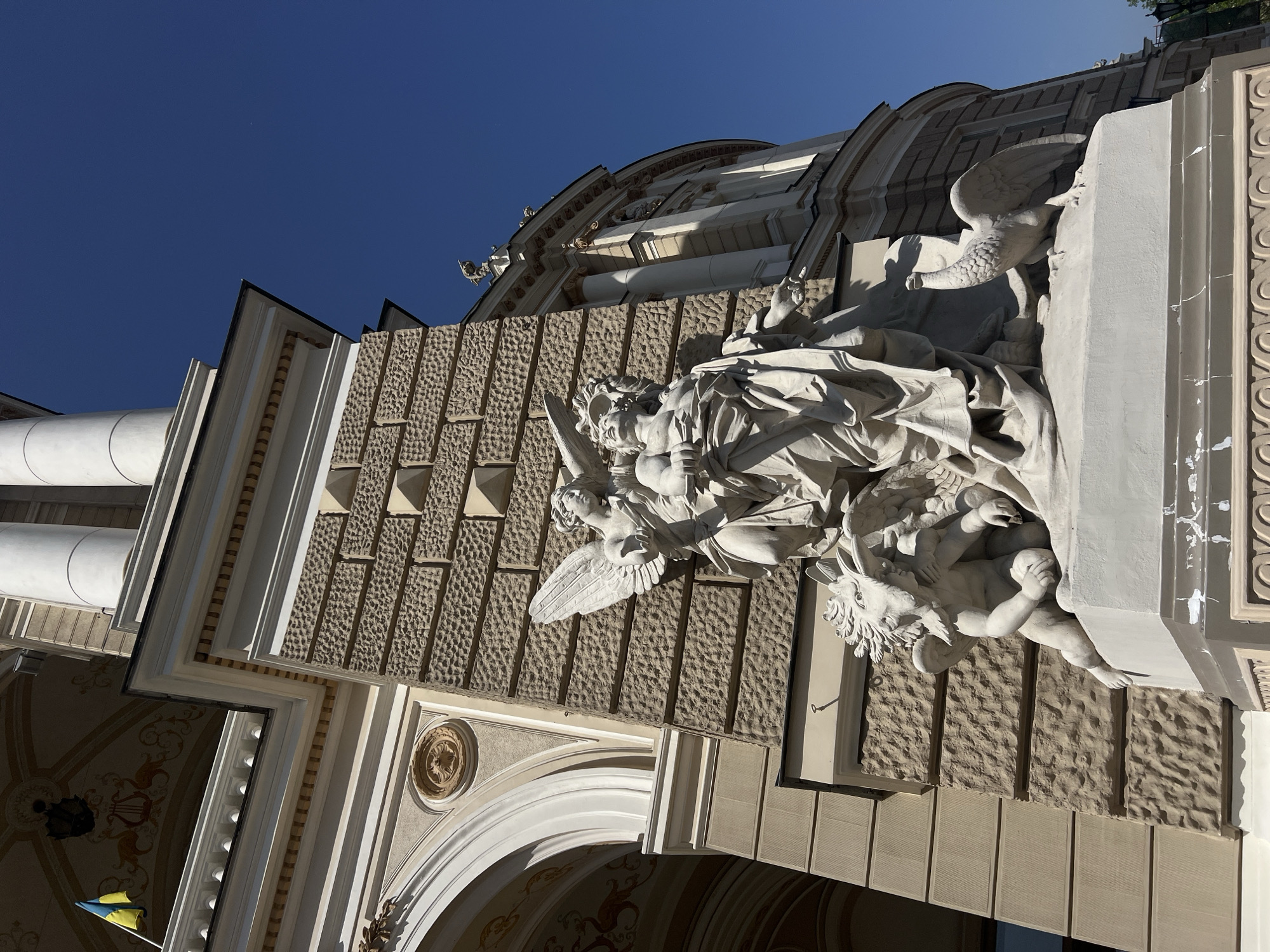 A low-angle, slightly tilted view of architectural details and sculptures on the exterior of the Odesa Opera and Ballet Theatre in Odesa, Ukraine. The perspective is from below, looking upwards, emphasizing the building's ornamentation against a clear blue sky.
The image focuses on a section of the building adorned with textured stone blocks, and elaborate white sculptures featuring human figures, angels, and possibly mythical creatures. A prominent sculpture of an angel with spread wings is visible on the facade. Other decorative elements include arched doorways, intricate molding, and cylindrical columns on the building's lower section.
In the bottom left corner, a small Ukrainian flag is discernible. The overall impression is one of classical architecture, rich in detail and sculptural adornment.