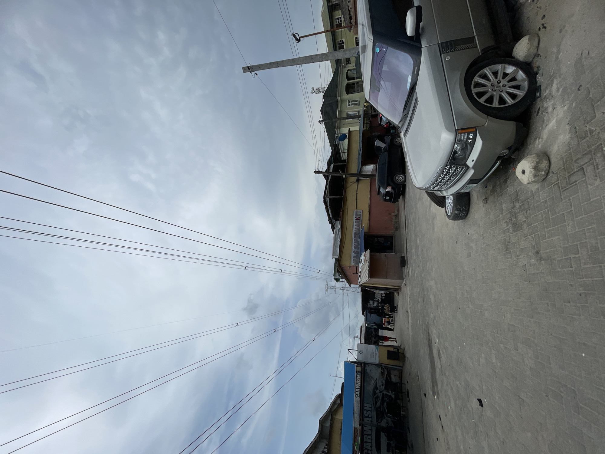 The image depicts a street in an urban environment. A light-colored Range Rover is parked on a brick-paved street, with two stones acting as wheel chocks. Power lines stretch across the sky above buildings of varying heights and colors. A "CARWASH" sign is visible on a building. People are visible further down the street, engaged in unspecified activities. The sky is overcast with clouds. The city name is unknown.
