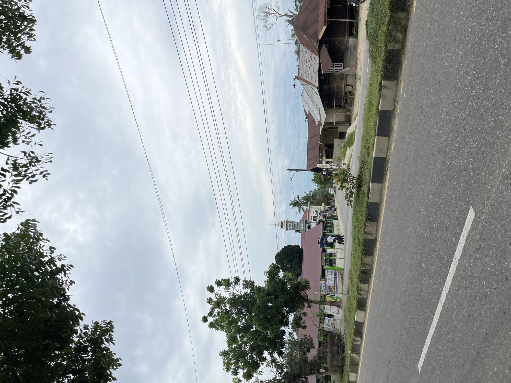 A medium shot captures a street scene in Tanahterban, Indonesia, with a cloudy sky overhead. In the foreground, a paved road with a white dashed line runs diagonally from the bottom left. A concrete curb with alternating black and white sections and a strip of grass separates the road from the sidewalk area. Along the sidewalk, there are several buildings. On the left, a low-rise building with a red roof is visible, possibly a school or community center, with posters displayed on its green fence. A tall, leafy tree stands in front of this building. Further down the street, a minaret topped with a dome and crescent moon rises above other buildings, indicating a mosque. To the right of the mosque, a row of simpler wooden structures with corrugated metal roofs are present. Several power lines crisscross the sky. A motorcycle with two riders is seen on the road in front of the buildings, and a car is visible in the distance on the left. The overall atmosphere appears to be a typical day in a semi-urban or rural Indonesian town.