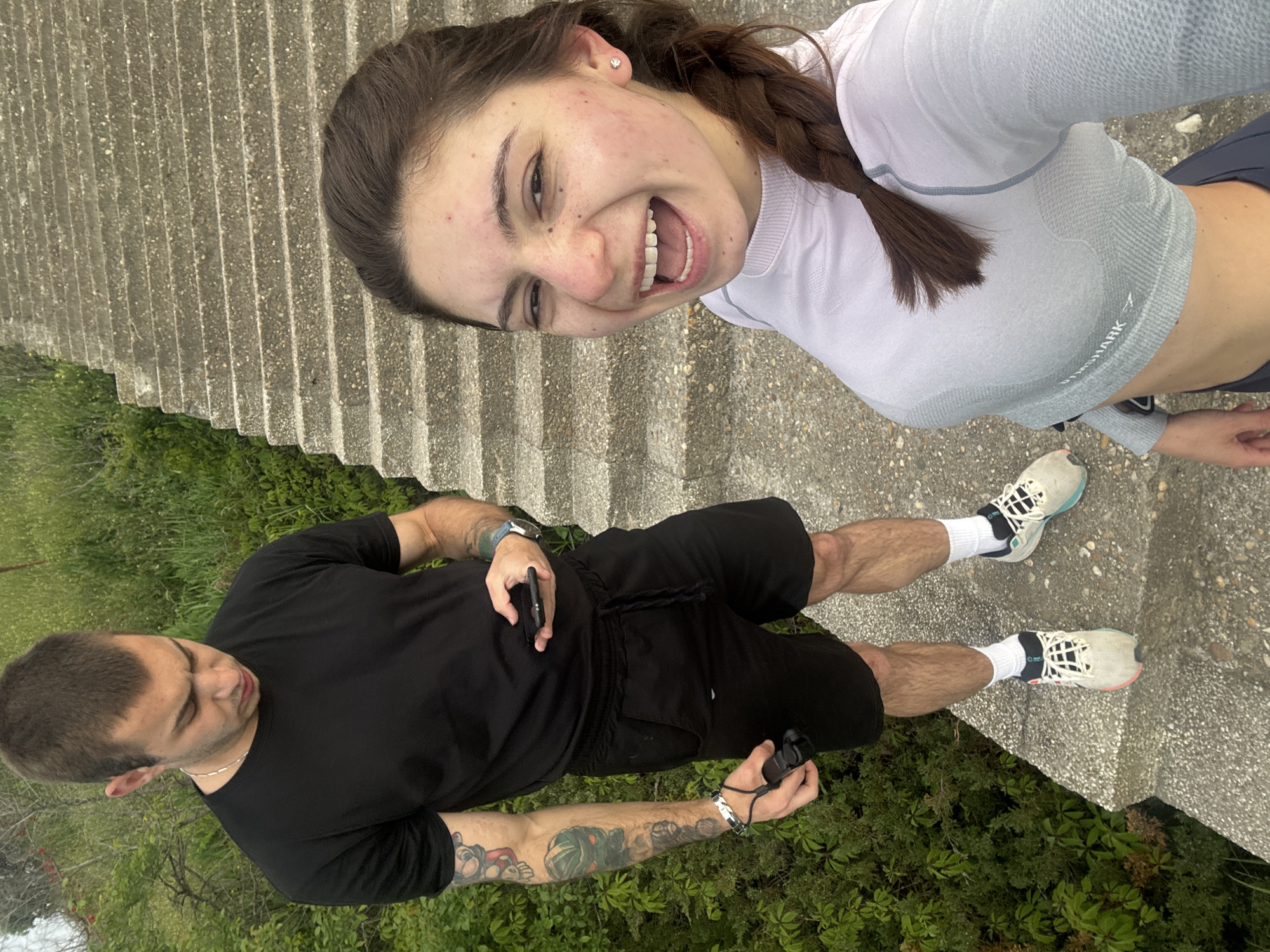 A photo taken in Varna, Bulgaria, likely on or near the Sea Garden stairs (due to the type of steps and surrounding vegetation). Two people are in the frame. A woman with brown hair in a braid, wearing a light-colored cropped athletic shirt and dark athletic leggings, is smiling and looking towards the camera, positioned above. A man wearing a black t-shirt and black shorts is standing below her. He has a shaved head and visible tattoos on his arm. He is holding a phone in one hand and what appears to be a camera or another device in the other. Both are standing next to concrete stairs and green vegetation is visible in the background.
