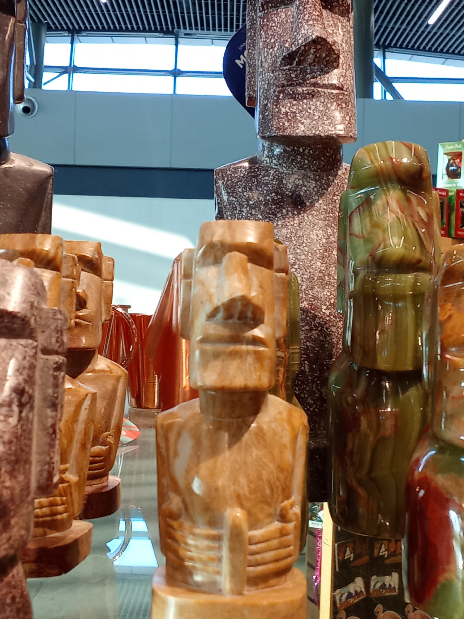 The image displays a collection of Moai statues, carved from various types of stone, arranged on a glass surface. The statues are predominantly in shades of brown, tan, and green, with intricate detailing on their faces and bodies. Some of the statues are larger than others, creating a tiered effect. In the background, a modern ceiling with visible lighting fixtures and a large window with a grid pattern can be seen. The overall environment suggests an indoor setting, possibly a shop or an exhibition space. There are no people visible in the image. The specific location in Pudahuel, Chile, is not evident from the visual information provided in the image.