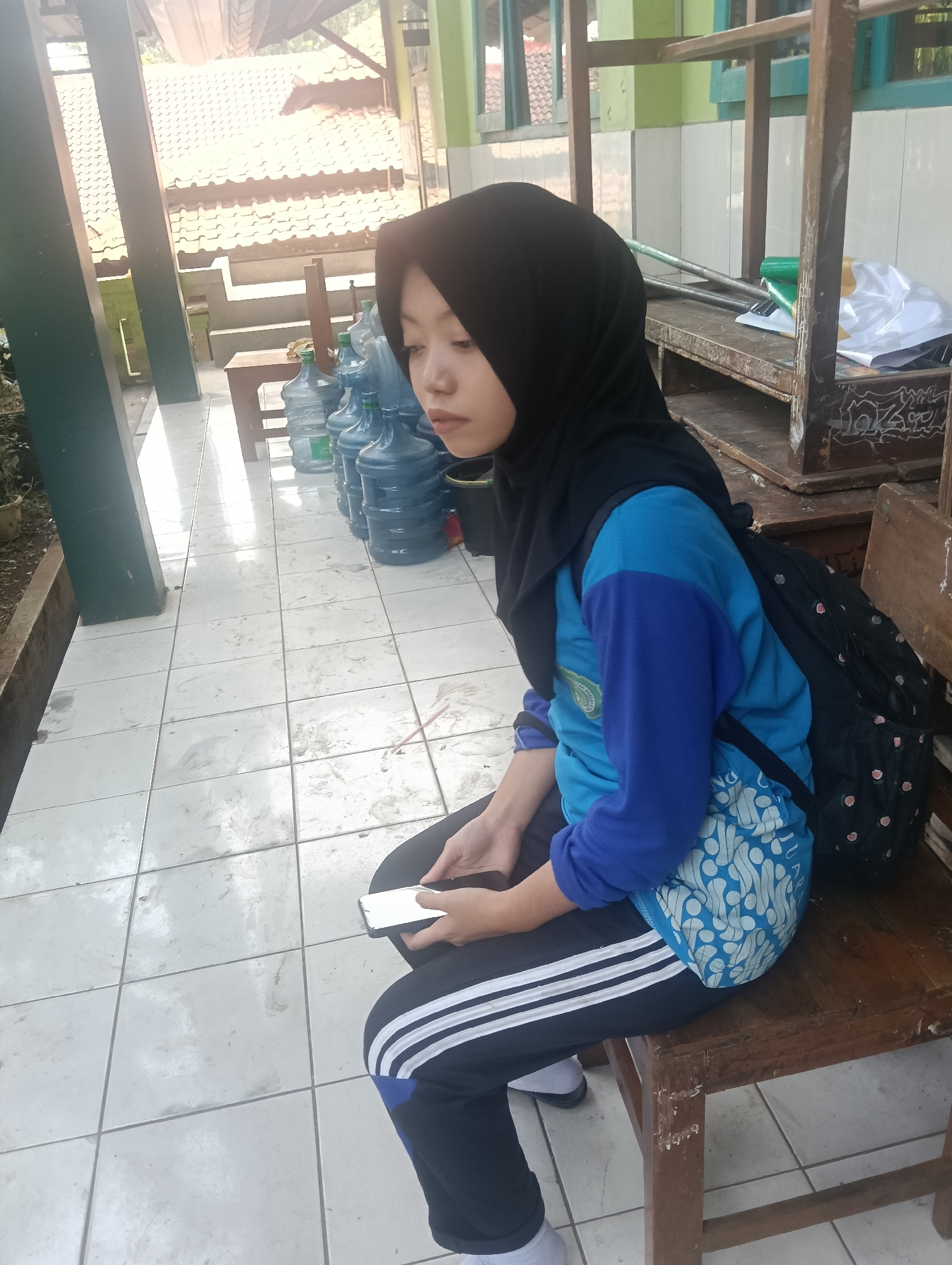 A young woman wearing a black hijab and a blue long-sleeved shirt with a blue patterned bag on her back sits on a wooden bench outdoors. She is looking down at her phone, which she holds in her hands. The bench is situated on a tiled floor next to a row of large water cooler bottles. In the background, there is a structure with a corrugated roof and some greenery. The scene appears to be in a school or community area, possibly located in Adinuso, Indonesia, given the context.
