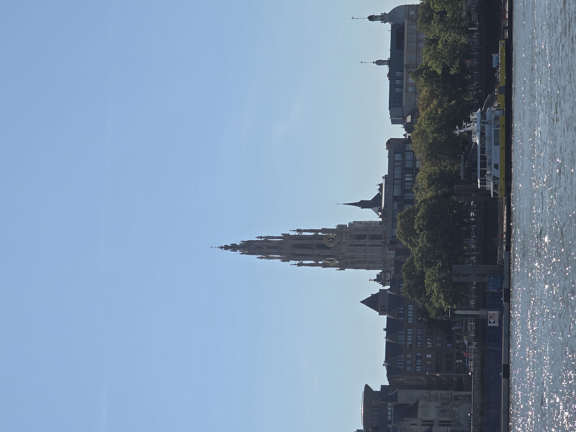 The image depicts a view of the Antwerp skyline from the Scheldt River. Dominating the view is the towering spire of the Cathedral of Our Lady, a Gothic cathedral situated in the city center. The cathedral's intricate architecture, including its clock faces, is clearly visible against the clear blue sky. Surrounding the cathedral are various other buildings, some modern and some with historical architectural styles, contributing to a dense urban landscape. In the foreground, the river is calm with its surface reflecting the sunlight, creating a shimmering effect. A white and yellow ferry or boat is moored along the riverbank, indicating a point of transit or tourism. Lush green trees line the riverbank between the water and the buildings, adding a natural element to the urban panorama. The overall scene suggests a bright, sunny day in Antwerp.