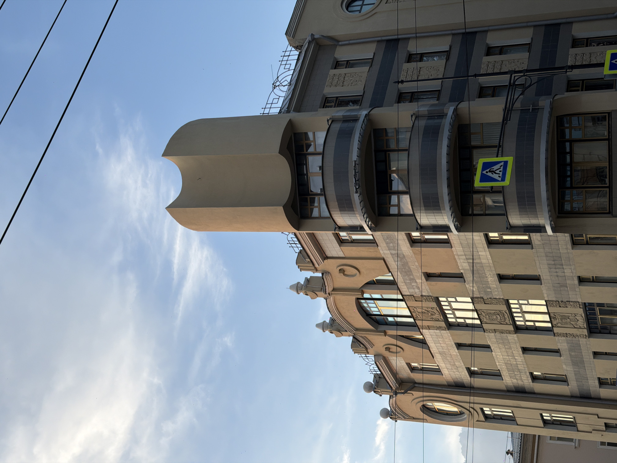 The image shows an Art Nouveau building on a clear day. The building is light beige with dark gray accents and features ornate decorative elements, including arched windows with decorative spires and carved reliefs. There are curved balconies with a dark railing. A distinctive rounded tower with a curved top rises from the side of the building. A yellow pedestrian crossing sign with a blue border is mounted on one of the balconies. Overhead, thick black power lines are visible against a pale blue sky with wispy clouds. The building appears to be located in a city.
This building is the former residential house of the merchant G.A. Kopylov, located at 11/13 Komsomolsky Prospekt in Perm, Russia.
