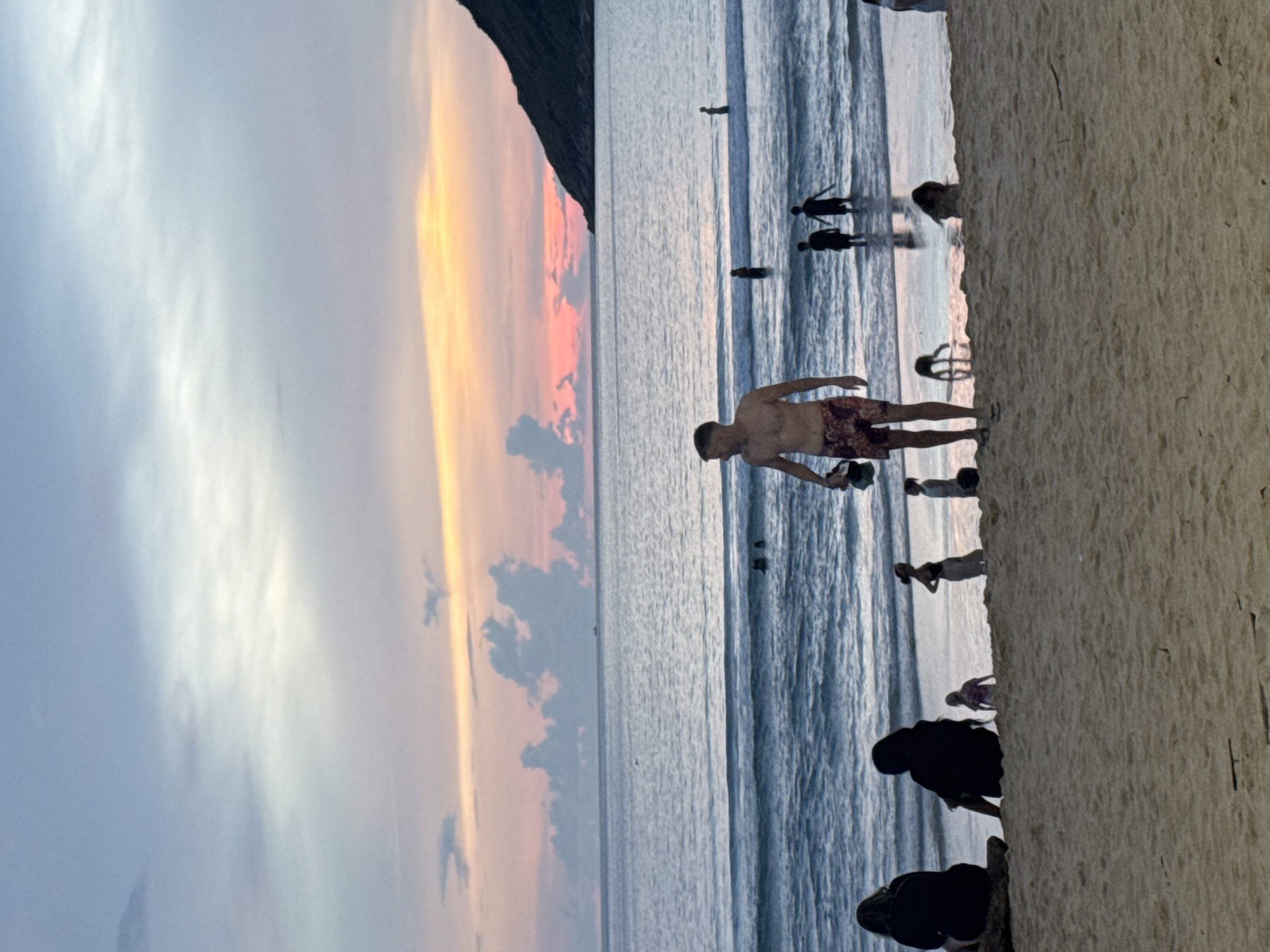 In the city of Unknown, Unknown, a twilight beach scene unfolds. The sky displays a gradient of soft blues and pinks, with wispy clouds and a faint orange glow near the horizon. The ocean, a calm expanse of blue water, gently laps against a sandy shore where several individuals are present.
In the foreground, a man in patterned swim trunks stands facing left, holding a dark object in his right hand. To his left, two figures sit silhouetted on a piece of driftwood, their backs to the camera. Further down the beach, a child in a pink garment stands near the water's edge, while other figures, including a woman in a black outfit and a child in a white top, are also visible.
In the middle ground, several people are wading in the shallow water, their figures dark against the brighter sea. Two figures, likely children, stand near the shoreline. In the background, a dark, tree-covered hill slopes down towards the ocean on the right side of the frame. The overall atmosphere is peaceful and serene, capturing a moment of leisure at the end of the day.