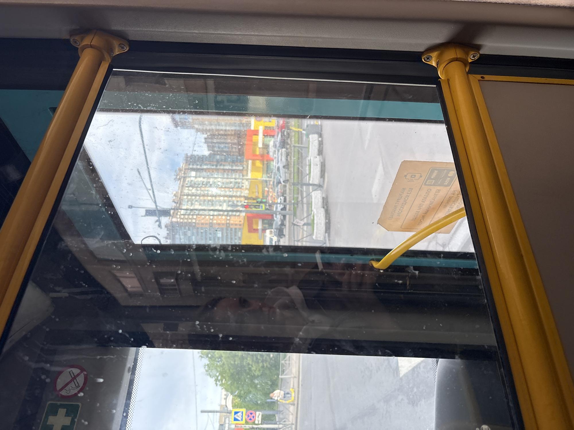 The image shows a view from inside a bus in Sinyavino, Russia. Outside the window are apartment buildings and street signs. A yellow handrail is visible in the foreground. A reflection of a person can be seen in the window.