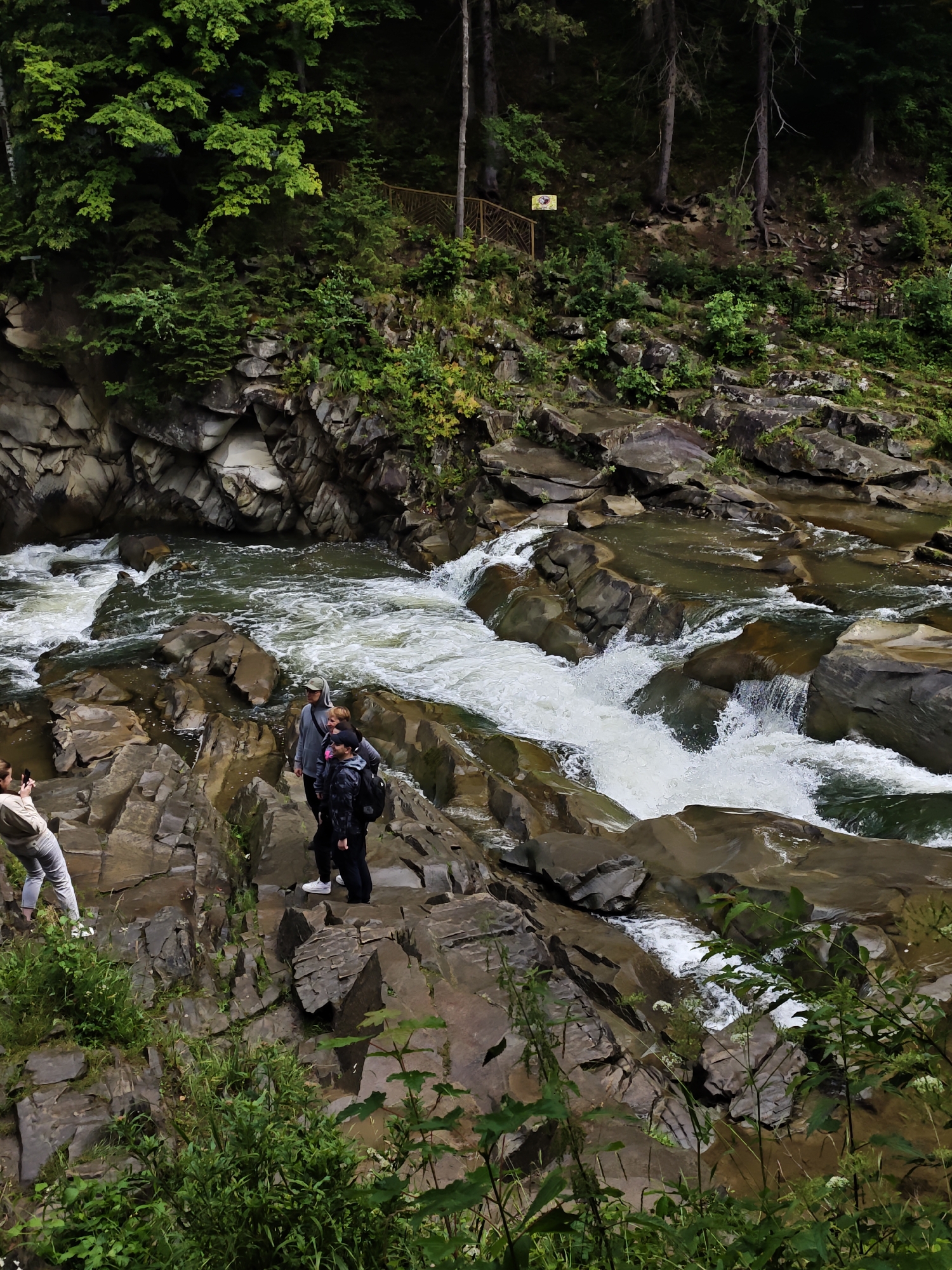 In Yaremche, Ukraine, a group of four individuals is captured in a photograph set against the backdrop of a rocky riverbed and lush forest. The scene is dominated by a rushing river, its white water cascading over numerous large, dark rocks. The surrounding environment is characterized by steep, rocky cliffs covered in green vegetation and trees.
On the left side of the frame, a person is standing on a rocky ledge, facing away from the camera and appearing to take a photograph with a phone. To their right, slightly further into the scene and closer to the river, three other individuals are positioned on a rocky outcrop. Two of them are standing close together, with one wearing a dark jacket and backpack, and the other in a lighter-colored hooded jacket. The third individual, who appears to be younger, stands slightly behind them. All three are facing towards the river.
The upper portion of the image reveals a densely forested hillside with tall trees and thick green foliage. A wooden railing and a yellow sign are visible on the hillside, suggesting a developed pathway or viewing area above the river. The overall atmosphere is one of natural beauty and outdoor recreation.