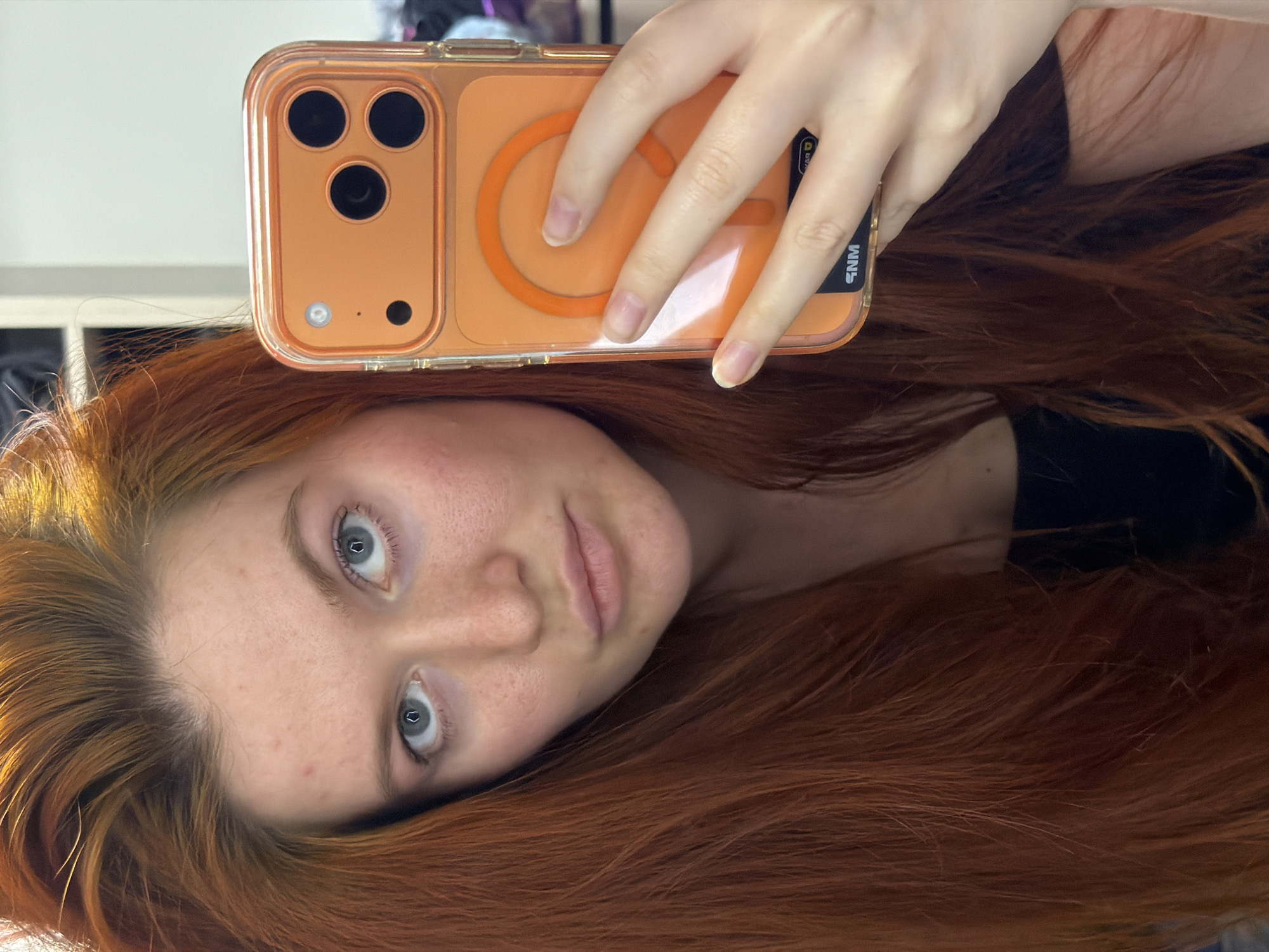A young woman with long, auburn hair is taking a selfie with her iPhone. She is wearing a black top. The phone is in an orange case with a magnetic ring. Her skin has freckles and some redness. The background appears to be indoors, with a slightly blurred closet or shelf visible behind her. There is no indication of the location being in Krasnodar, Russia.