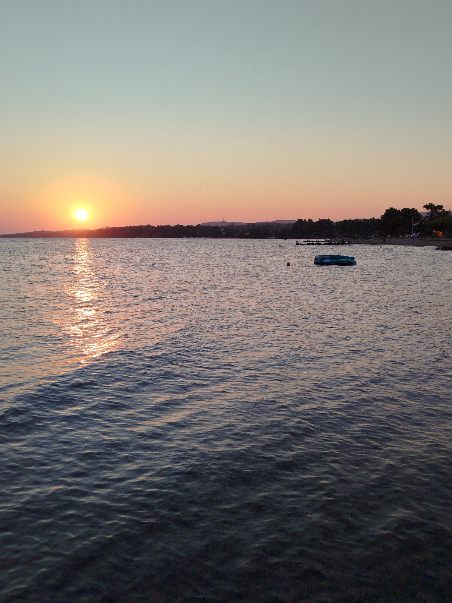 An outdoor, eye-level, vertical, wide shot captures the serene beauty of a tranquil sea at sunset. The sun, a vibrant orange orb, casts a shimmering golden path across the water's surface. In the distance, a tree-lined shore forms a dark silhouette against the pastel hues of the twilight sky, which transitions from a pale blue at the zenith to a soft orange and pink near the horizon. A small, dark, inflatable boat floats idly on the water, accompanied by a red buoy. The gentle waves create a rippling texture on the sea, reflecting the ambient light. The scene appears to be set in Sithonia Municipal Unit, Greece, a location known for its picturesque coastal landscapes.