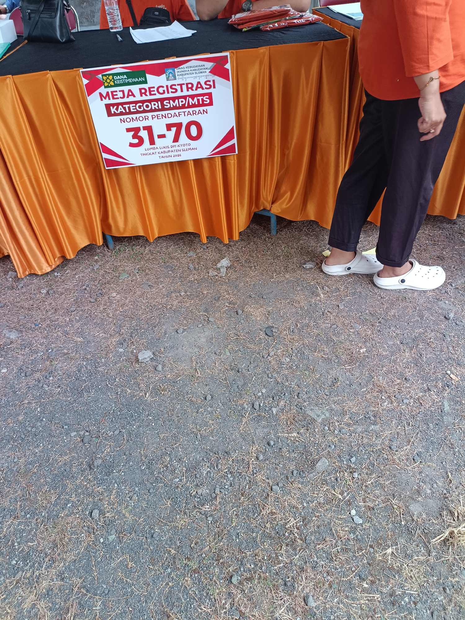 A woman wearing black pants and white clogs stands to the right of a table draped with an orange tablecloth. The table has a sign with red and white elements that reads "MEJA REGISTRASI KATEGORI SMP/MTS NOMOR PENDAFTARAN 31-70 LOMBA LUKIS DIY-KYOTO TINGKAT KABUPATEN SLEMAN TAHUN 2026." The ground is covered with gravel and dry grass.