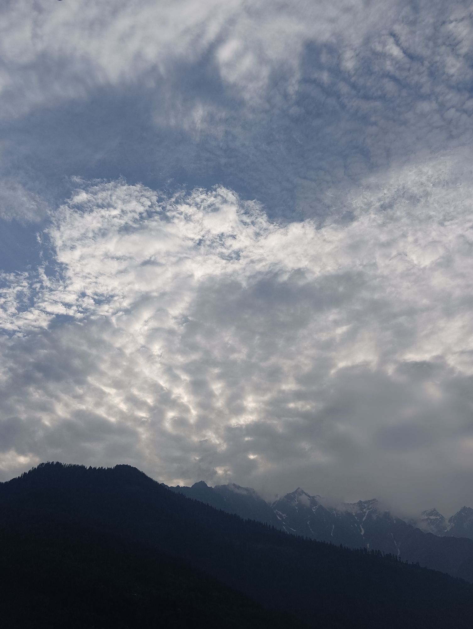 This image depicts a mountainous landscape with a dramatic sky. The foreground is dominated by the dark silhouette of a mountain range, dotted with trees along its ridge. Further in the distance, snow-capped peaks of other mountains emerge through a layer of clouds.
The sky is filled with a dynamic display of clouds. Towards the top of the frame, wispy, light-colored clouds are scattered against a pale blue sky. As the eye moves down, the clouds become denser and more textured, forming undulating patterns that suggest movement and varying depths. Some areas of the sky are brightly illuminated, hinting at the sun's position behind the clouds, while other parts are darker and more shadowed.
The overall impression is one of grandeur and natural beauty. The contrast between the dark mountains and the illuminated sky creates a sense of drama. The presence of snow on the distant peaks suggests a high-altitude location. Given the location context of Vashisht, India, this scene likely represents the Himalayas. The lighting and cloud cover suggest it is likely daytime, possibly late afternoon or early morning, when light conditions can be more varied and dramatic. There is no visible text in the image.