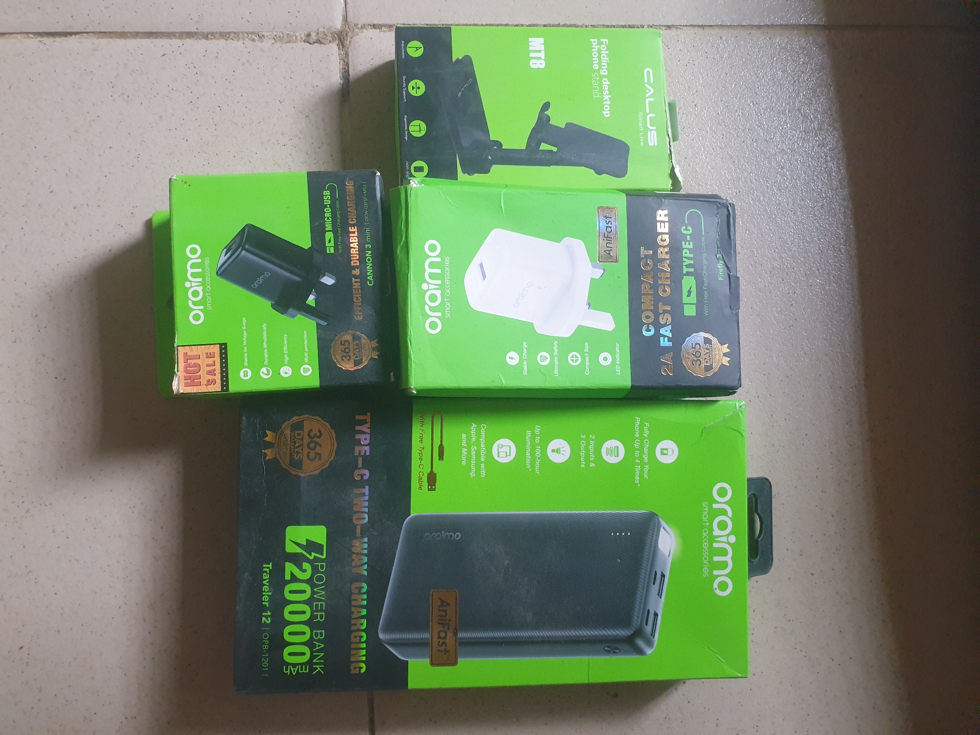 The image displays a collection of electronic accessories, primarily Orimo branded, arranged on a light-colored, textured surface that appears to be a floor or wall. The scene is well-lit, suggesting it's likely daytime indoors.
The main subjects are the product packaging for various Orimo smart accessories. These include:
* **Orimo Smart Accessories Power Bank:** A black power bank is partially visible, encased in its green packaging. The packaging highlights "TYPE-C TWO-WAY CHARGING" and a capacity of "20000 mAh" with the model "Traveler 12 | OPB-1201". It also mentions "AniFast" charging and a "365 DAYS WARRANTY".
* **Orimo Smart Accessories Chargers:** Two green boxes are visible, one for a "COMPACT 2A FAST CHARGER" with "TYPE-C" and "AniFast" branding, and another for a charger labeled "EFFICIENT & DURABLE CHARGING" with a "MICRO-USB" connector and "CANNON 3 mini" model. Both feature "365 DAYS WARRANTY".
* **Calus Folding Desktop Phone Stand:** A green box displays a "MT8" model, described as a "Folding desktop phone stand". It shows images of the stand in use and highlights features like "non-slip", "stable support", and "dynamic design".
Notable details include the prominent use of green packaging, the presence of "AniFast" and "365 DAYS WARRANTY" on multiple items, and clear labeling of product types and features. The overall impression is a collection of new, boxed electronic accessories, likely for sale or recently purchased, set against a simple, neutral background that doesn't provide specific location cues beyond suggesting an indoor setting. The text on the packaging is in English.