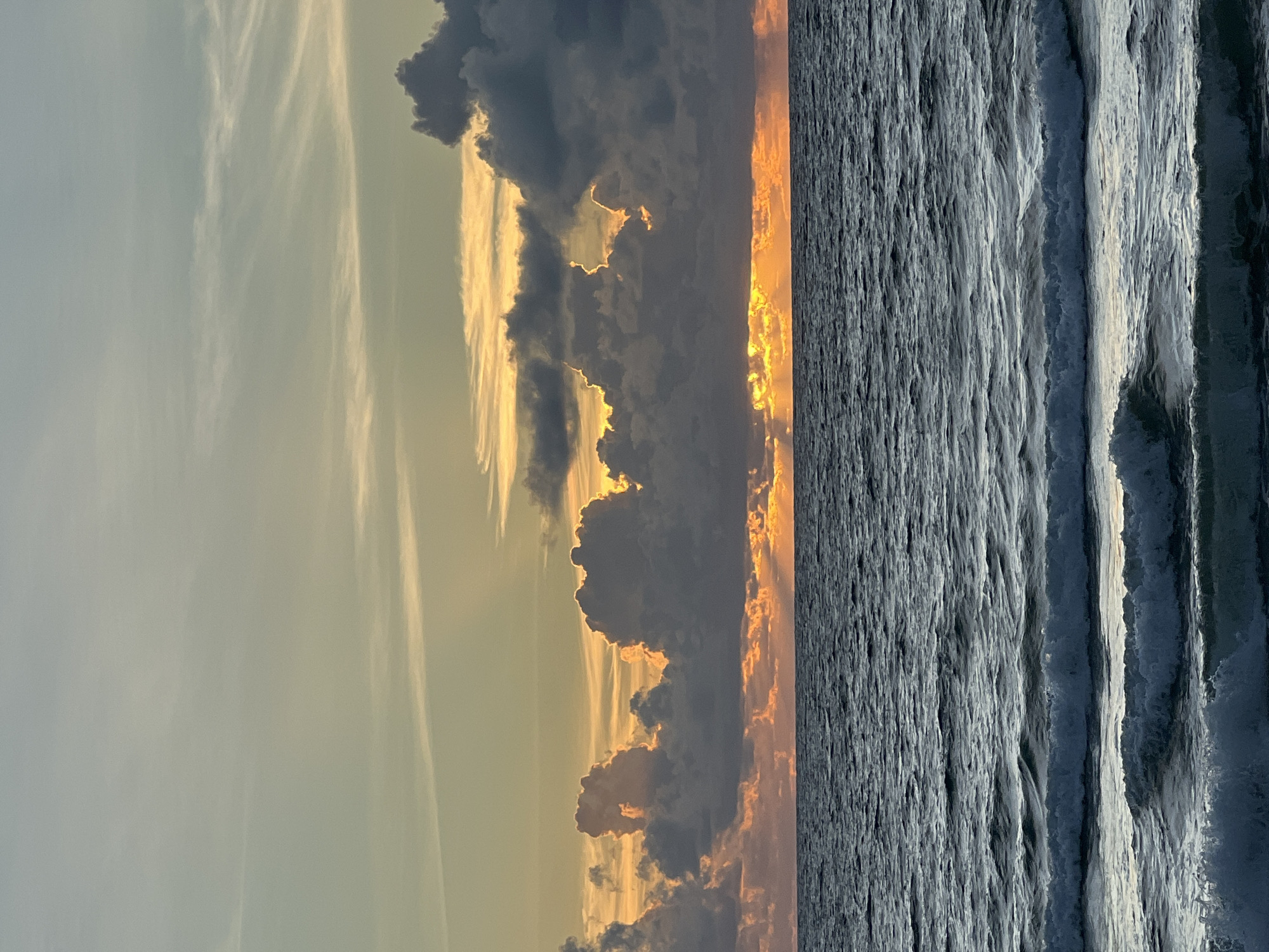 This is a photograph of a sunset over the ocean. The sky is filled with dramatic clouds, some of which are illuminated by the setting sun, creating streaks of orange and yellow light. The ocean below is dark and choppy, with waves breaking near the shore. The horizon line is visible, separating the sky from the sea. There are no people or man-made objects visible in the image. The scene appears to be natural and serene. The location is unknown.