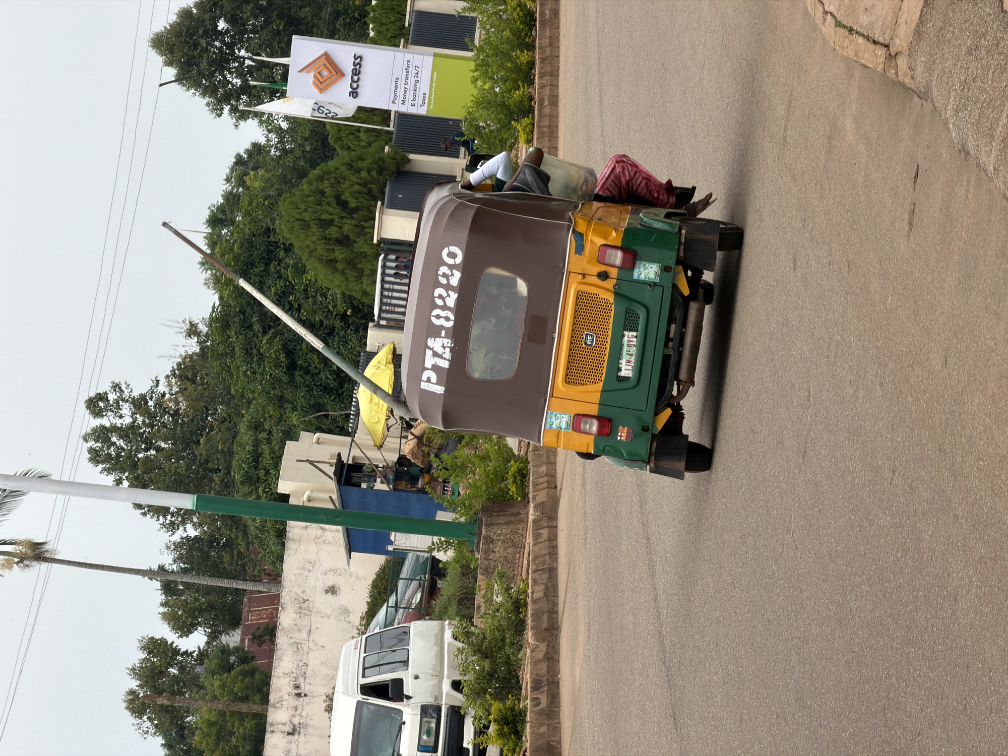 The image captures a tricycle, known as an "okada" or "keke," in motion on a paved road in Bukuru, Nigeria. The tricycle is predominantly brown and green, with yellow accents. It is carrying at least two passengers, one of whom is visible in the passenger seat wearing a red and white striped garment, and another person, possibly the driver, partially obscured. The tricycle has a license plate that reads "LJN 45RYG."
Behind the tricycle, a white panel van is parked. In the background, a sign with the "access" logo and the text "Payments," "Money transfers," "E banking 24/7," and "Taxes" is visible. The scene appears to be set during the daytime, under an overcast sky, suggesting a possibly humid climate typical of Nigeria. Lush green trees and vegetation frame the scene, indicating an outdoor environment. The overall impression is a glimpse into daily life and transportation in a Nigerian urban or semi-urban setting.