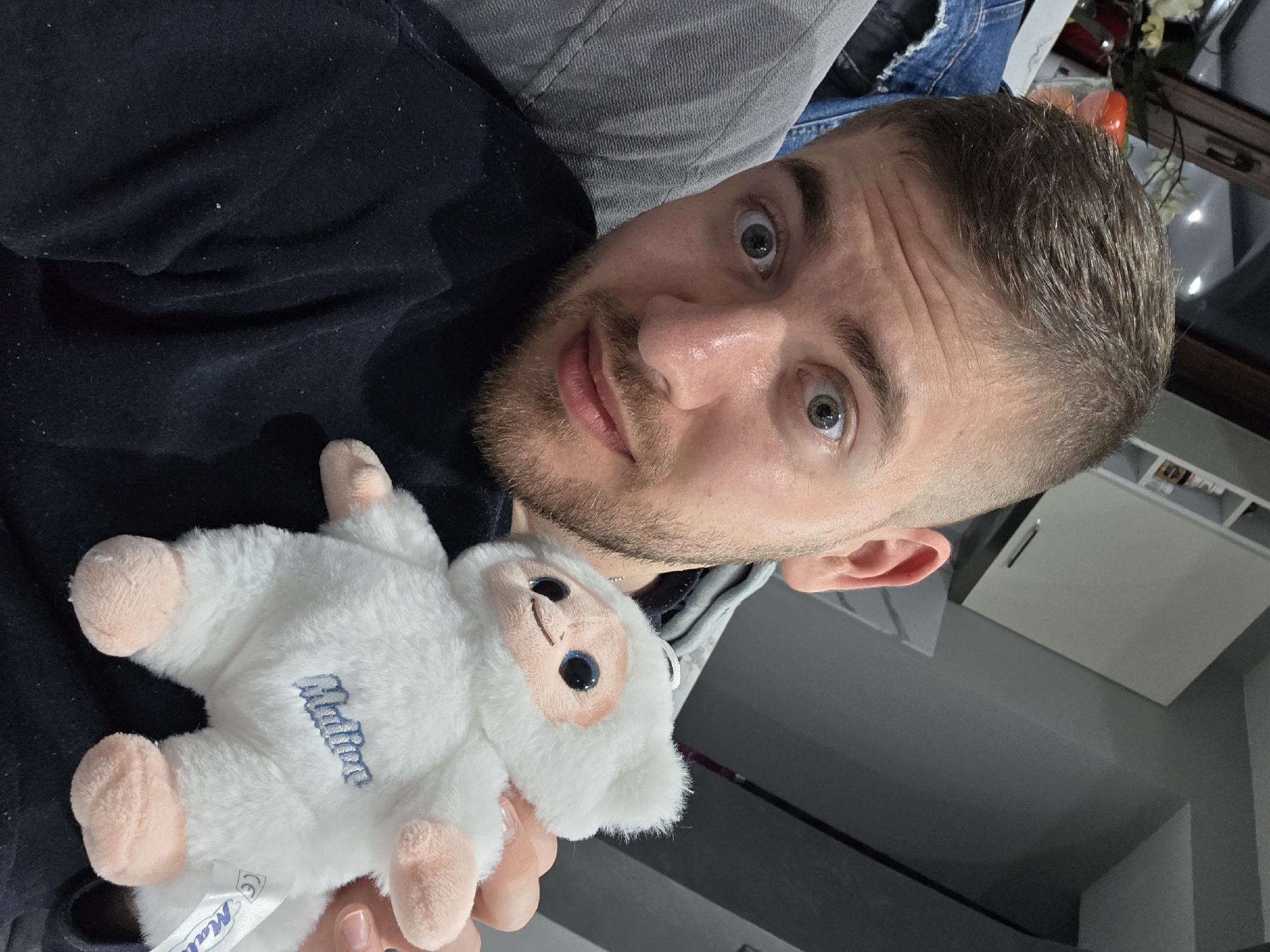 A man is holding a white stuffed animal. The man has short brown hair, light skin, a beard, and light-colored eyes. He is wearing a dark-colored shirt. The stuffed animal has pink paws and the word "Malina" embroidered on it. The environment is an interior space with a grey wall and furniture in the background. The location is not identifiable as Yagodovo, Bulgaria.